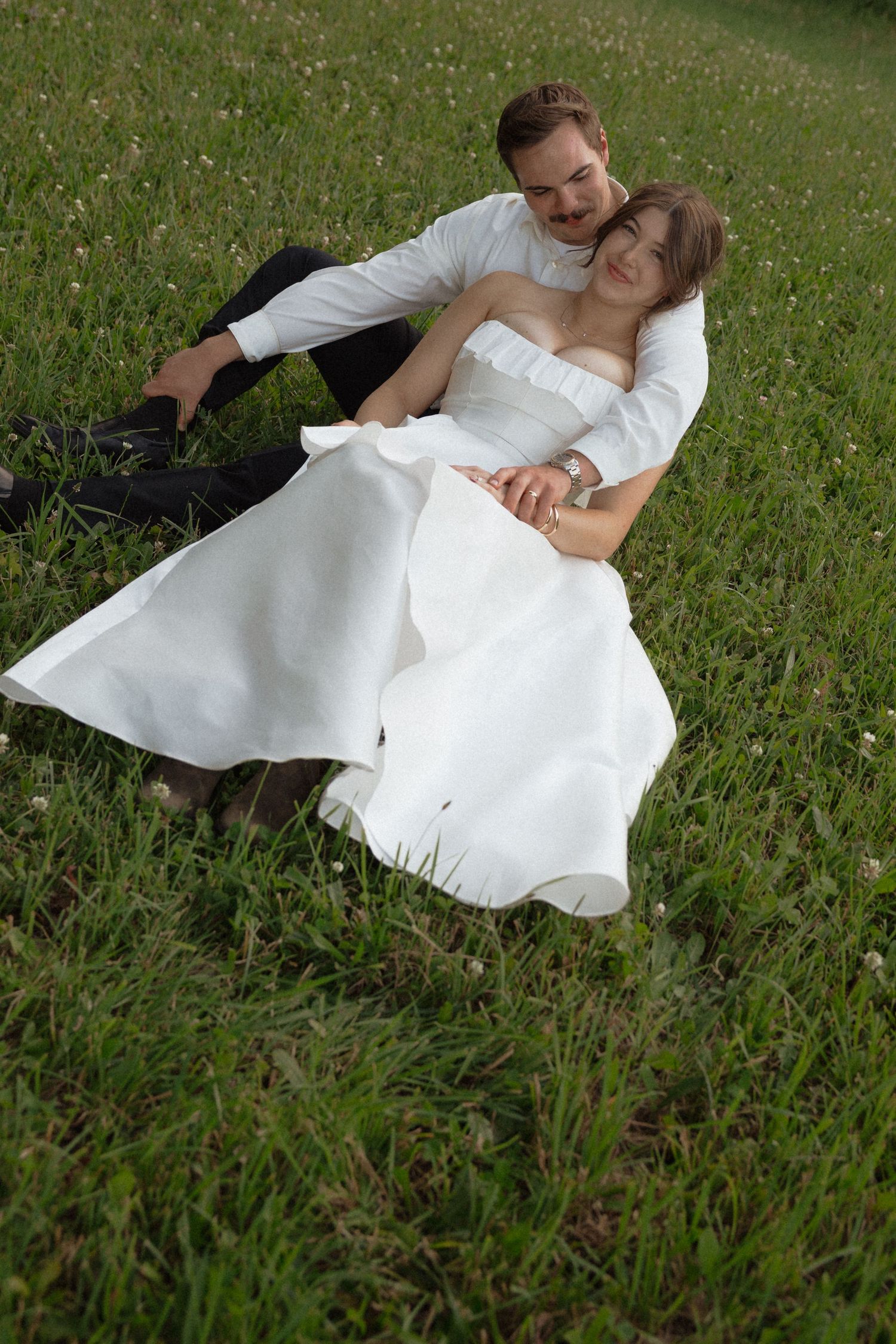 Bride and groom relax together in a green meadow, bride in strapless white gown leaning against groom.