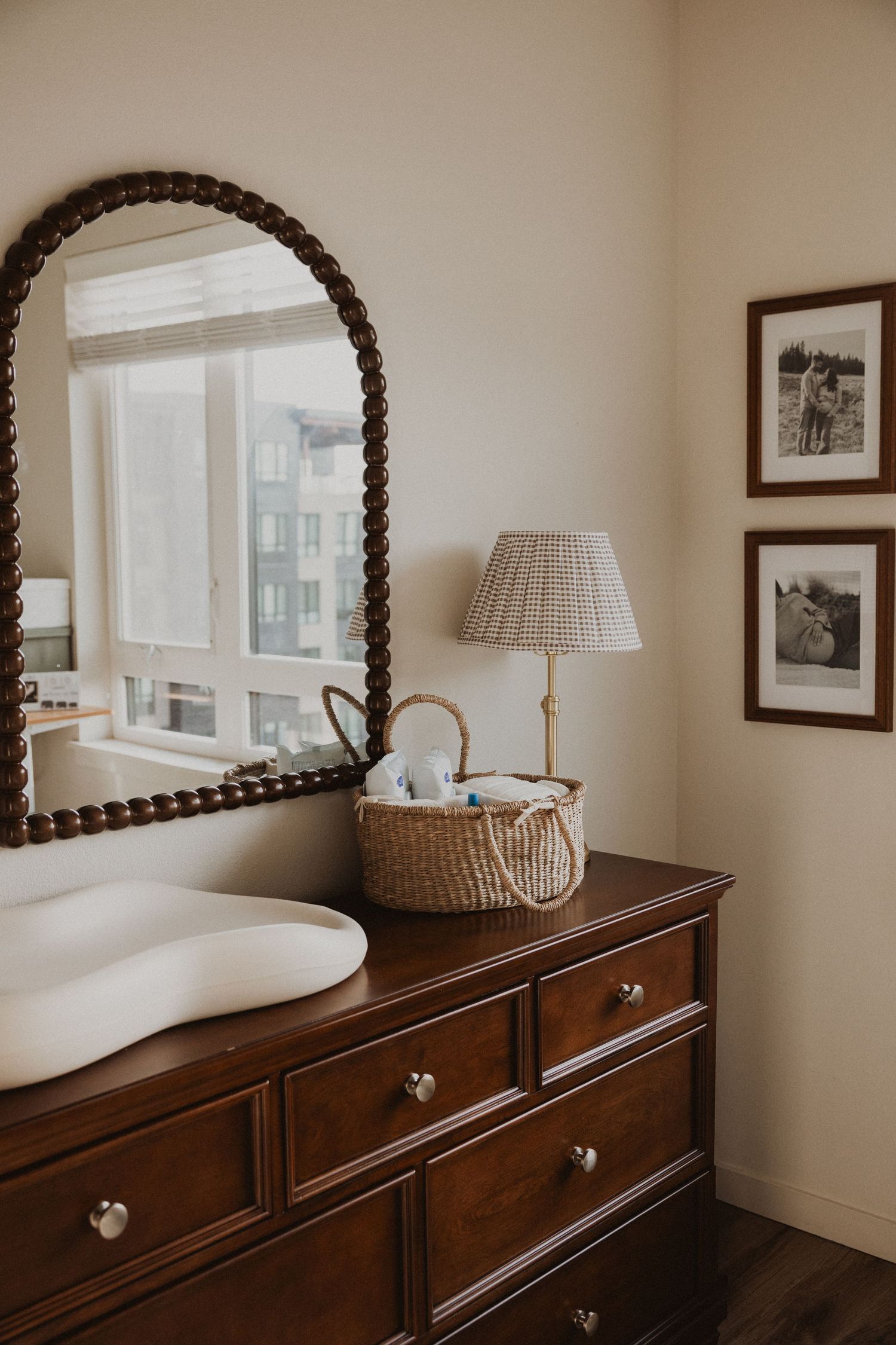 Dark wood dresser with arched beaded mirror, wicker basket, and brass lamp in a neutral nursery.