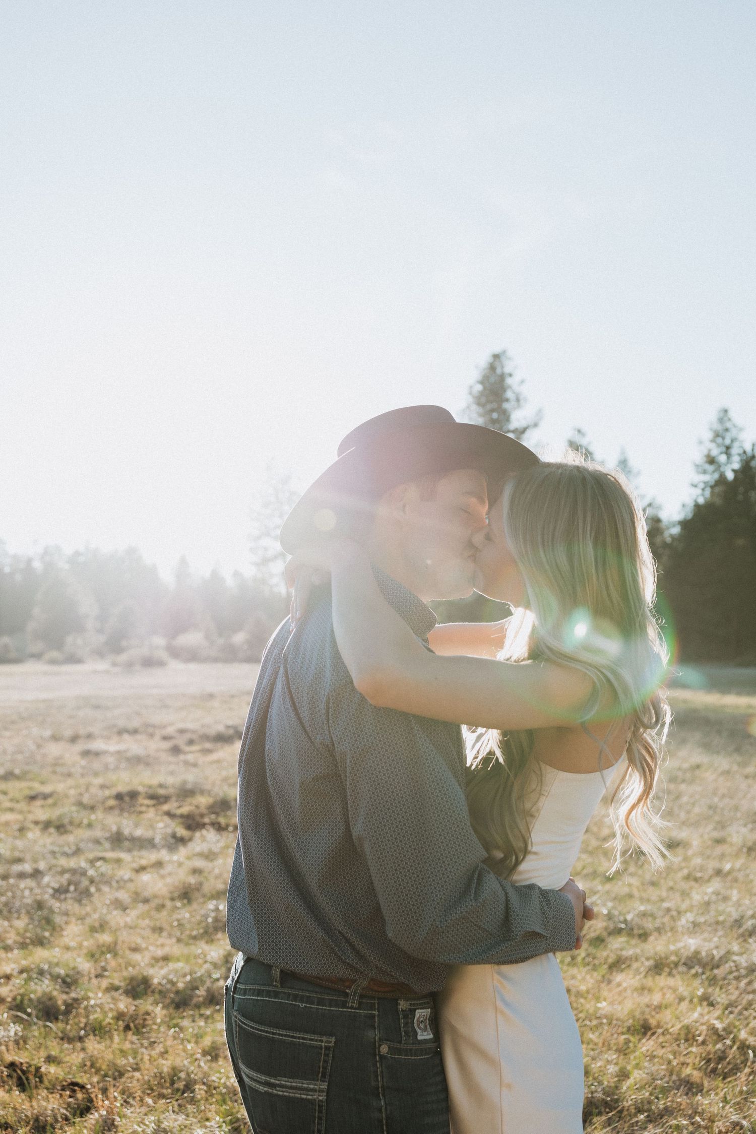 Couple sharing a romantic kiss in a sunlit field with lens flare, surrounded by trees and golden light.
