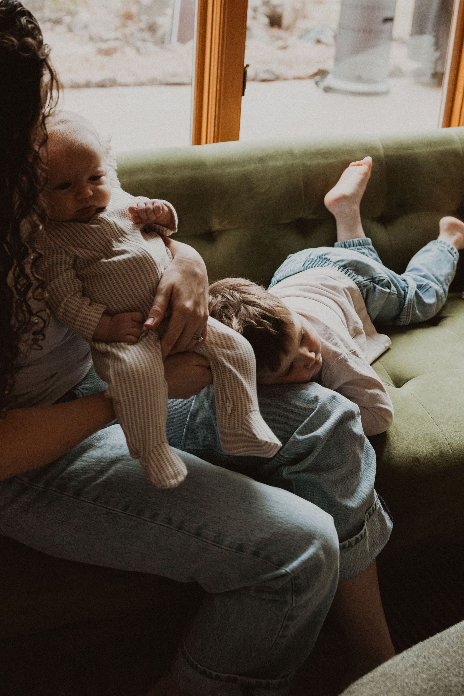 Mother holding newborn baby while toddler rests on her lap on green sofa.