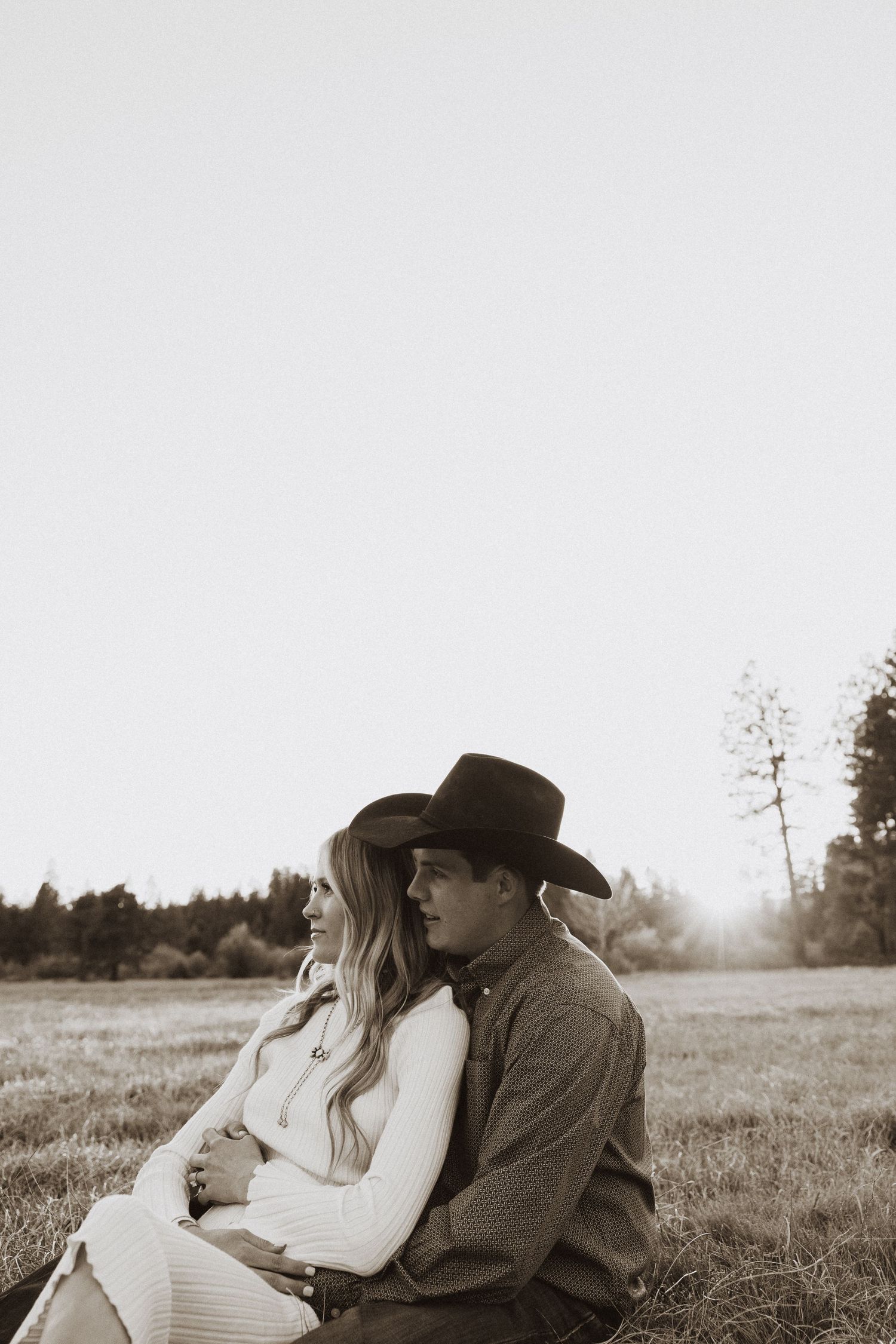 A couple sits together in a field at sunset, the man wearing a cowboy hat embracing a woman in a white dress.