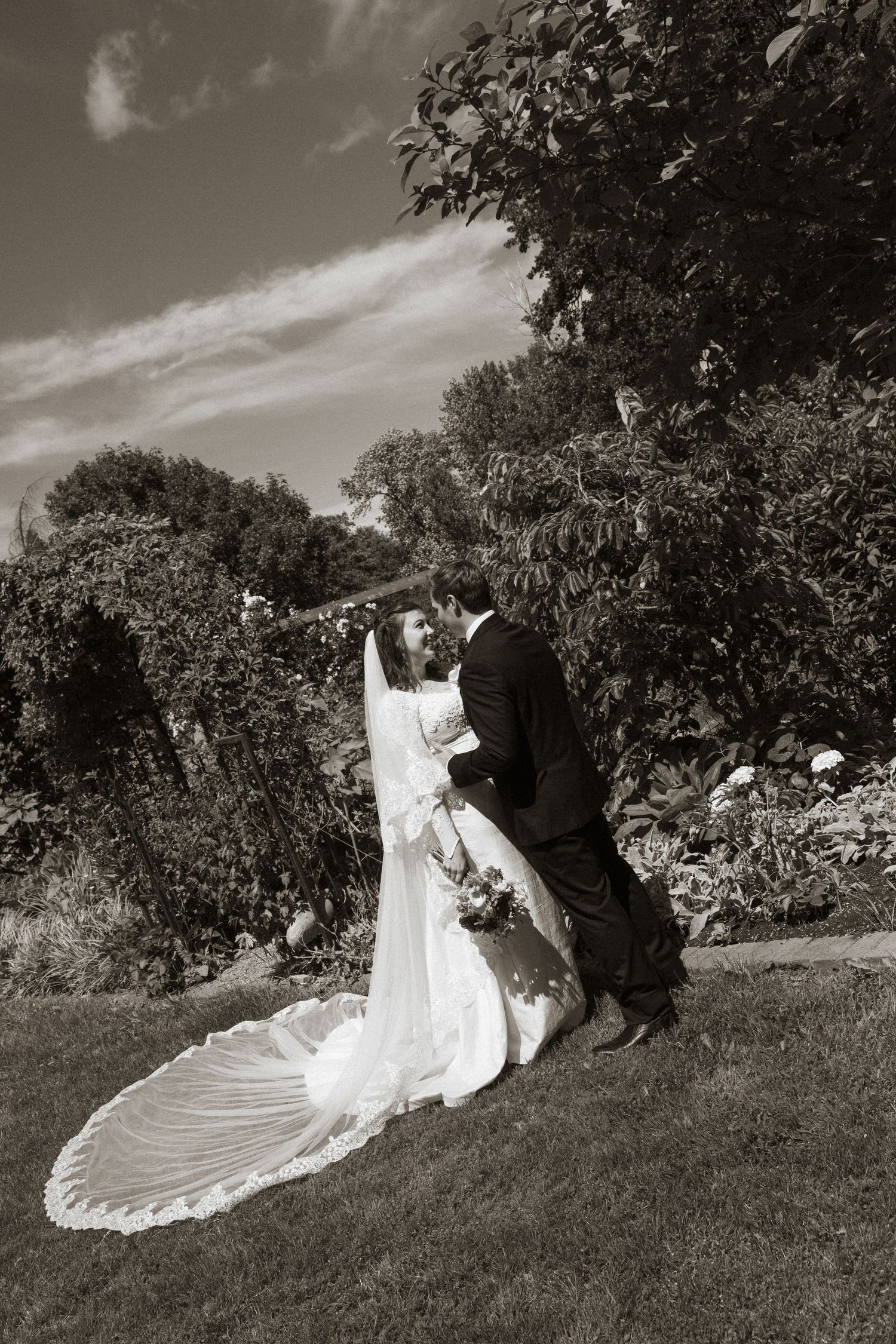 Black and white photo of a bride and groom kissing in a garden, brides long veil trailing behind.