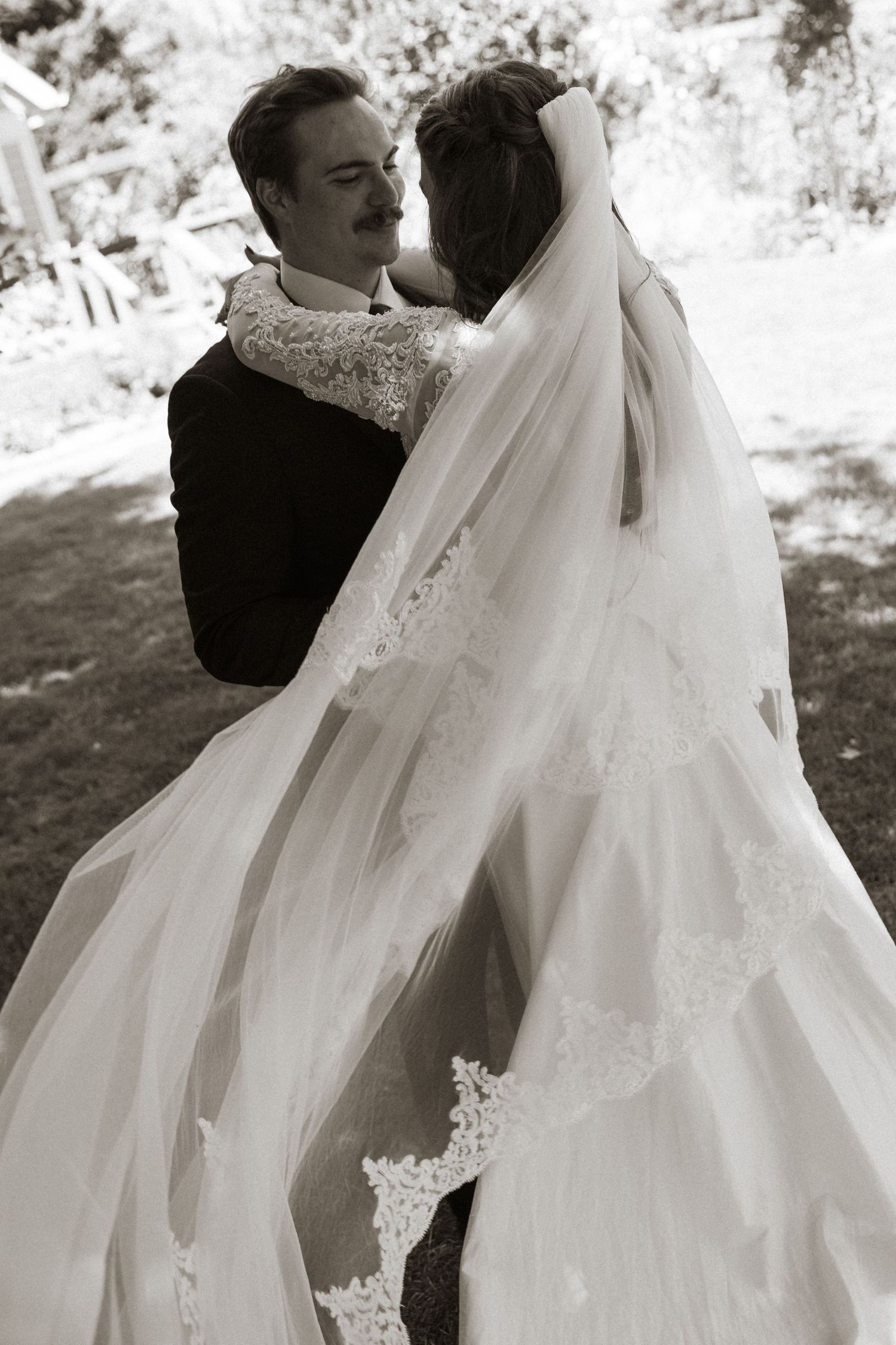 Bride and groom embrace romantically in black and white wedding photo outdoors.