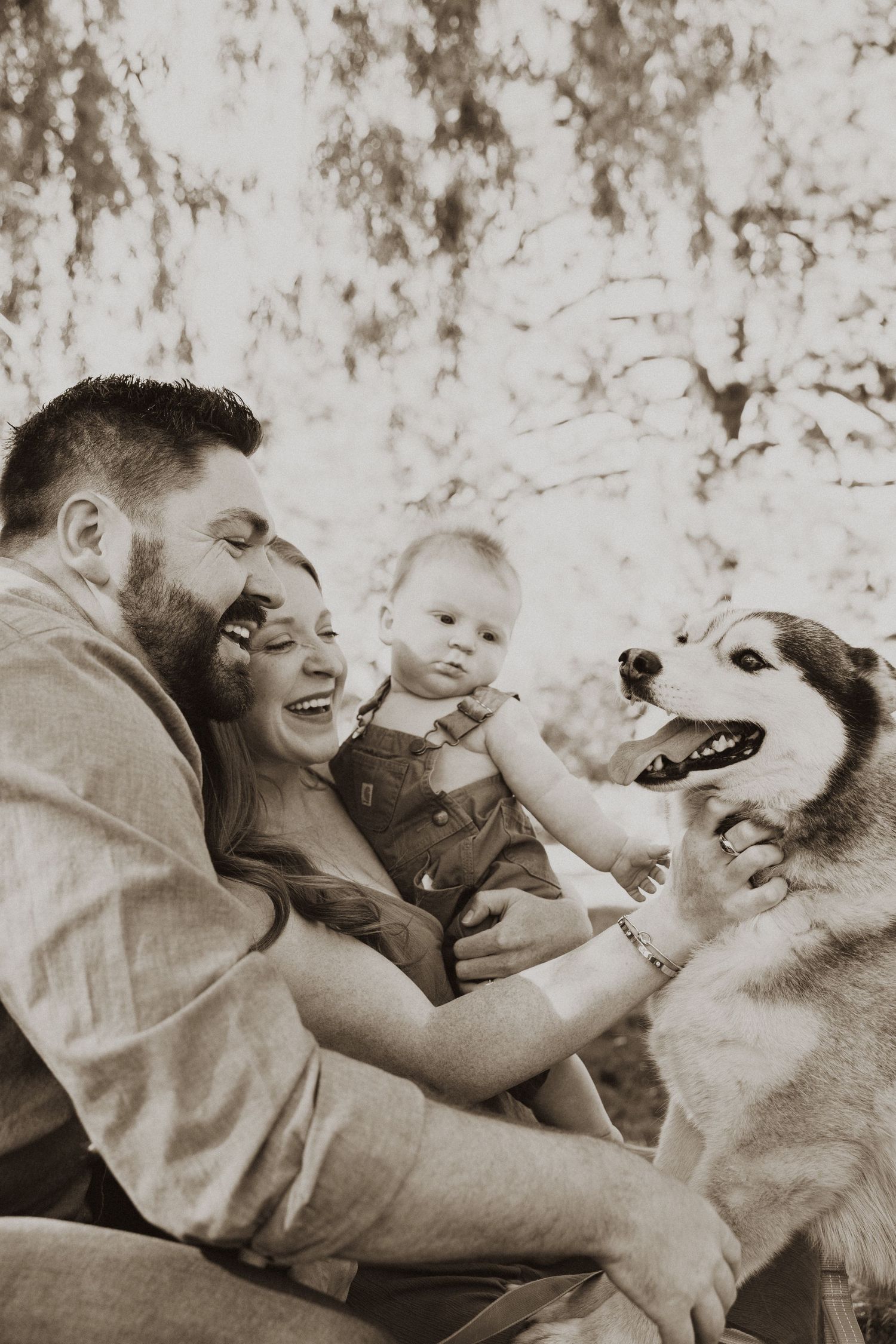 Sepia-toned family portrait with parents, baby, and Husky dog outdoors among trees.