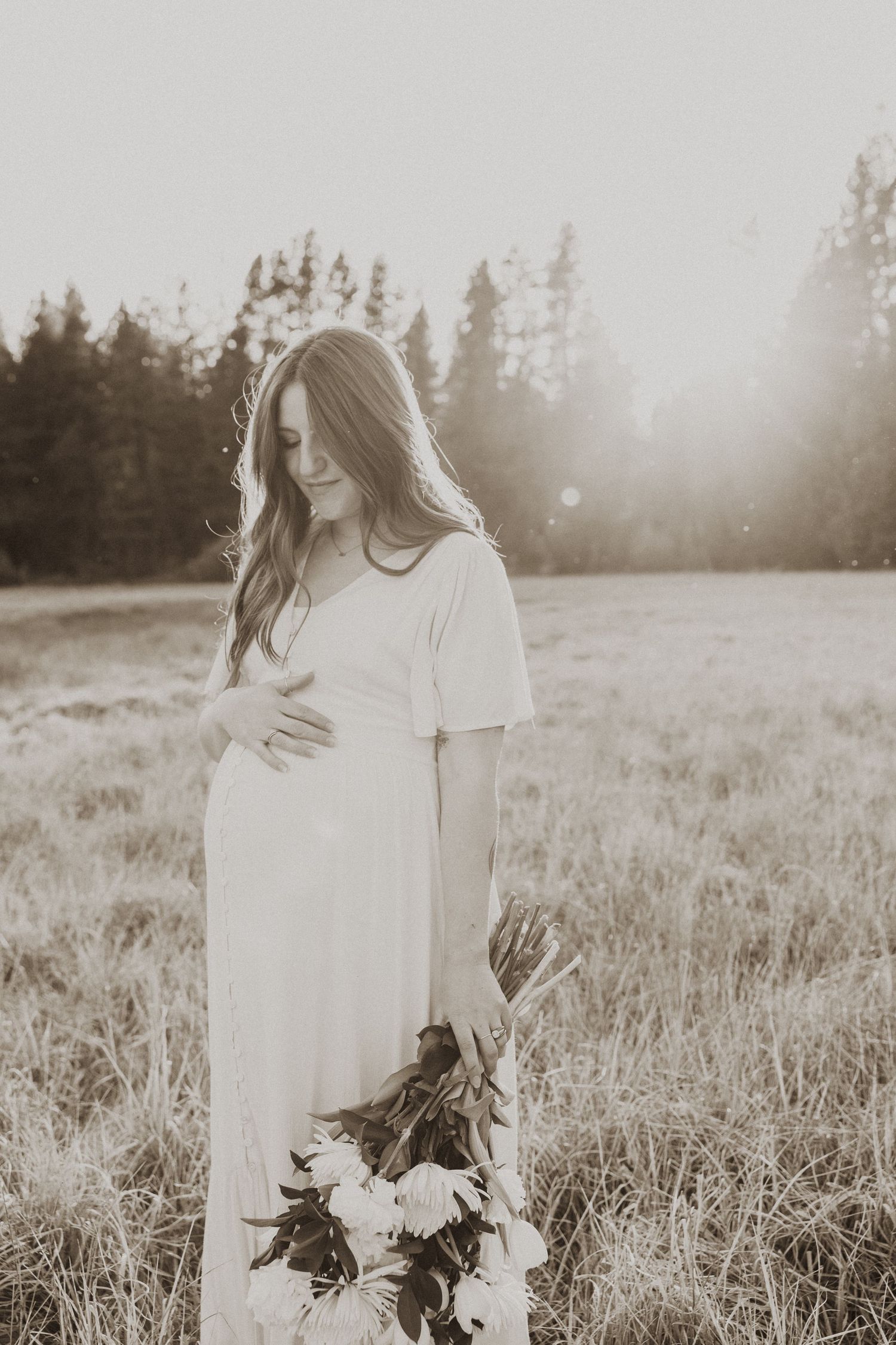 Pregnant woman in white dress holding flowers in sunlit field.
