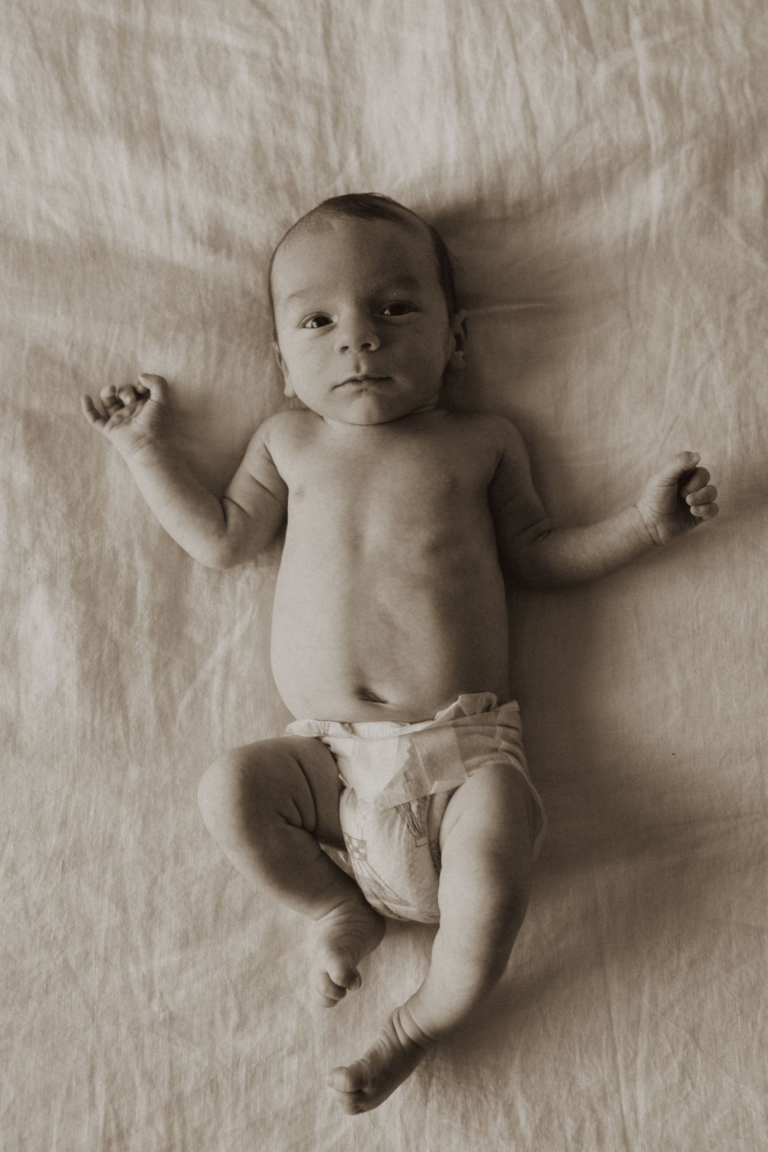 Sepia-toned newborn baby lying on back in diaper on soft blanket, arms raised, looking at camera.