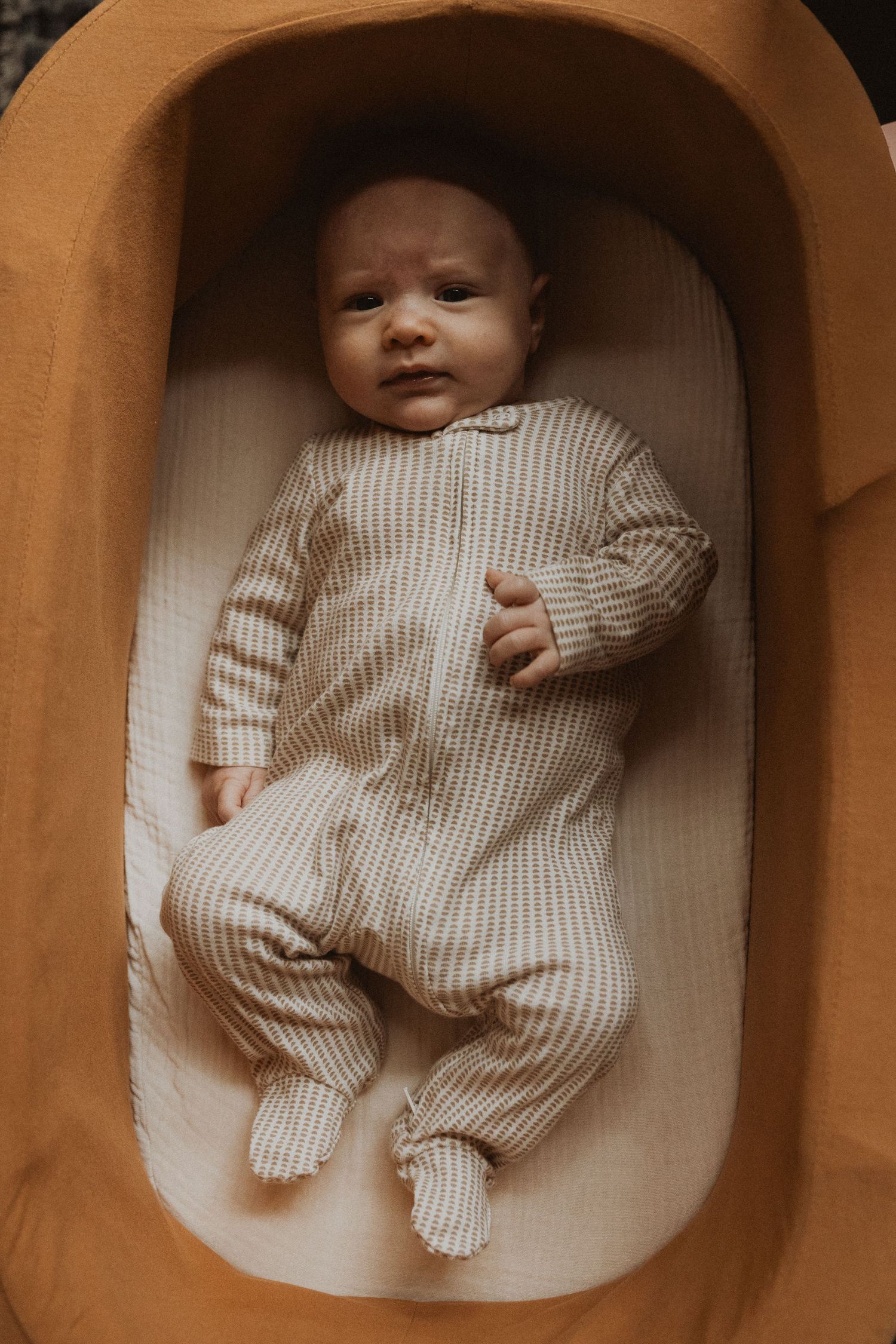 Peaceful newborn baby in striped onesie lying in a mustard yellow bassinet looking up.