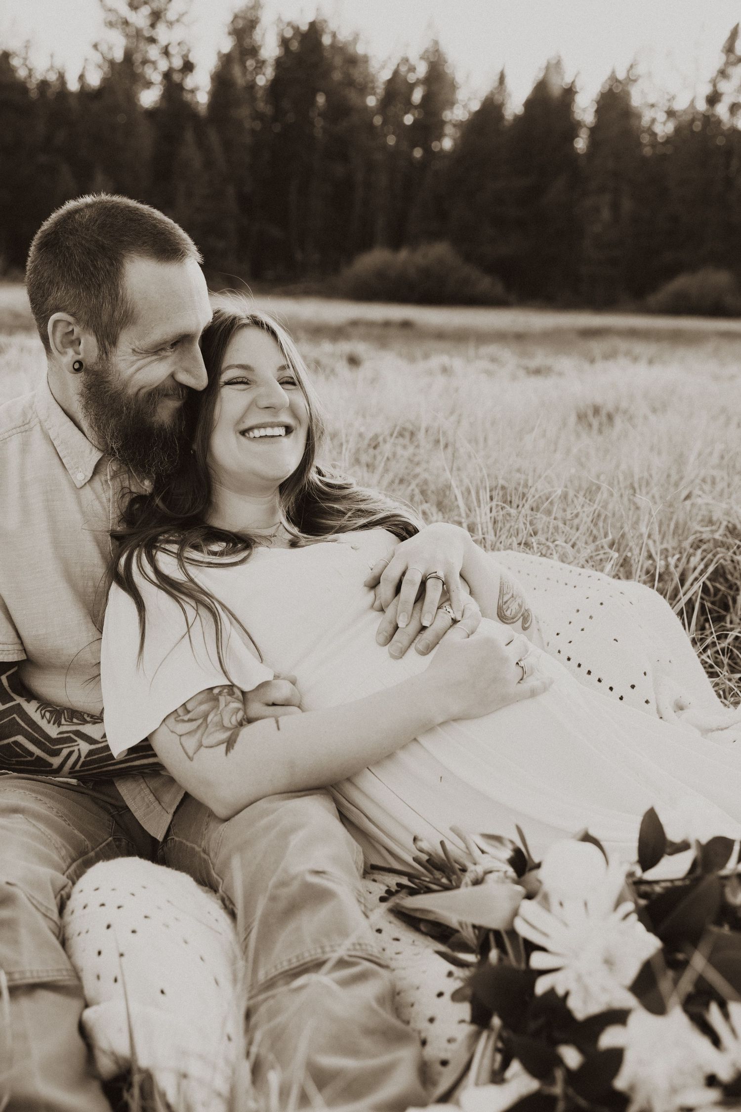 Happy couple sitting in a field, man embracing pregnant woman in sepia-toned maternity photo.