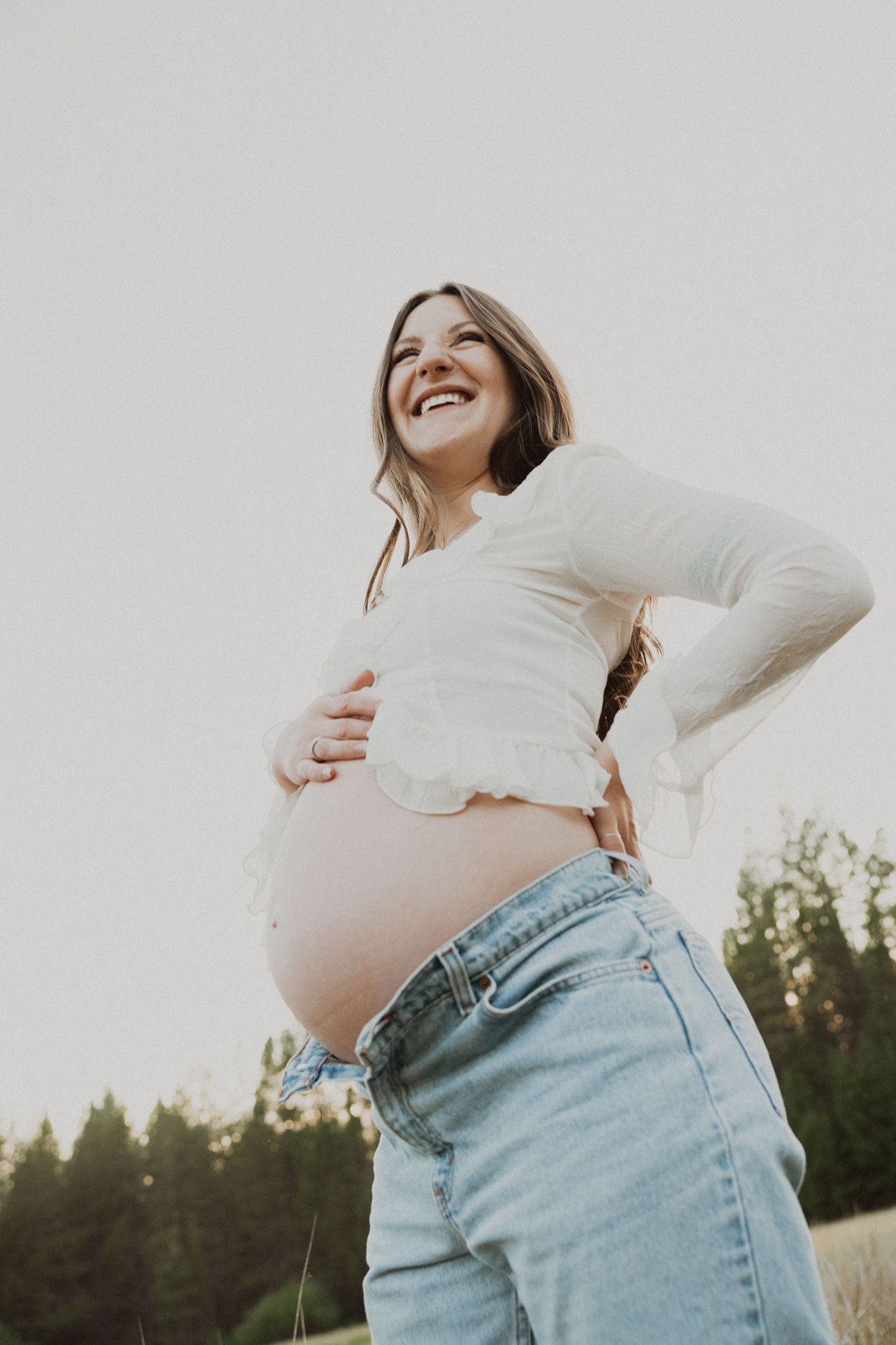 Smiling pregnant woman outdoors in white crop top and jeans, cradling her baby bump against a natural background.