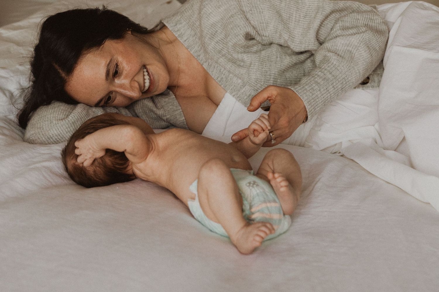 Smiling mother lying on white bed gazing lovingly at her newborn baby in a diaper during a lifestyle newborn session.