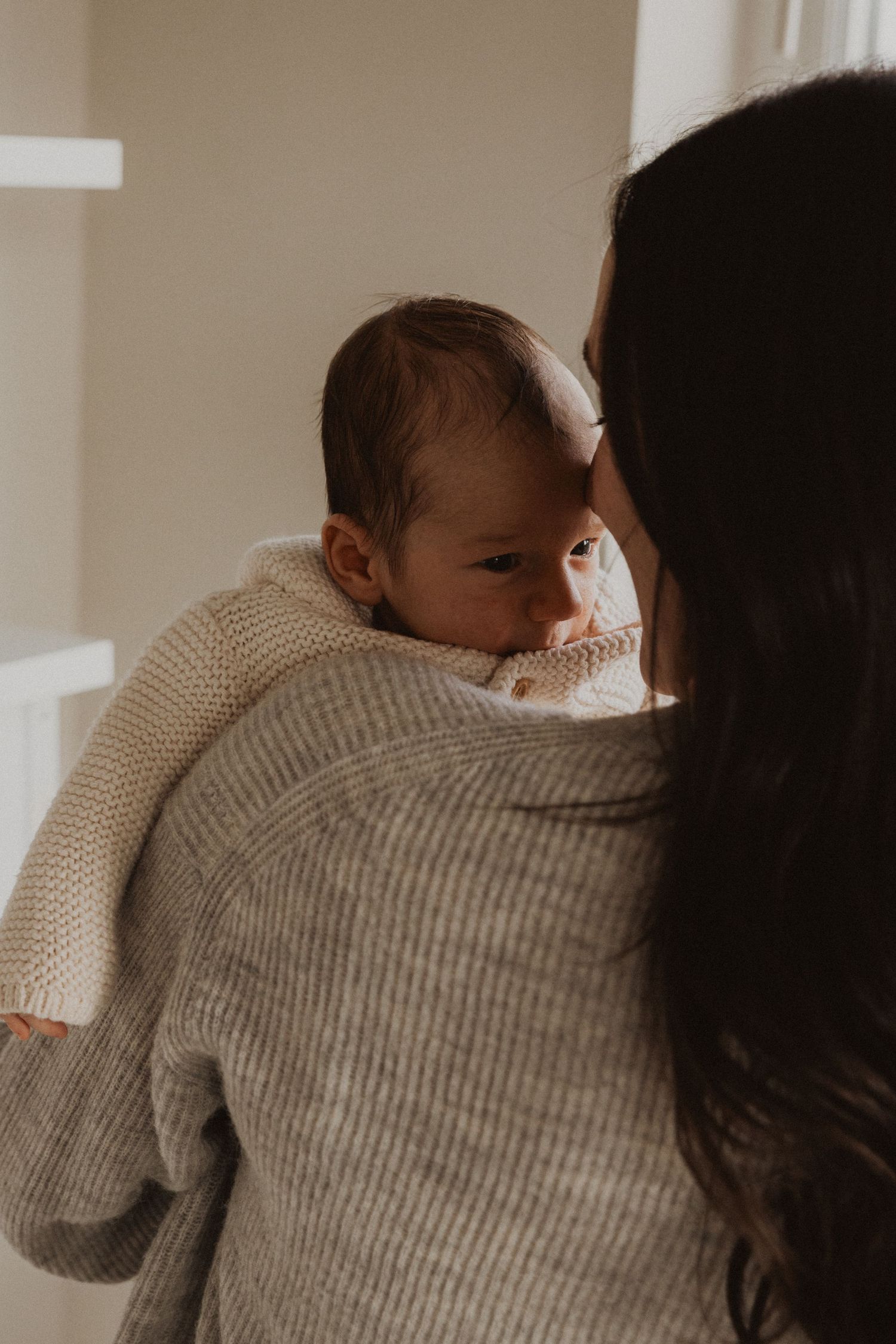 A mother in a gray sweater holds her newborn baby dressed in a cream knit outfit against her shoulder.