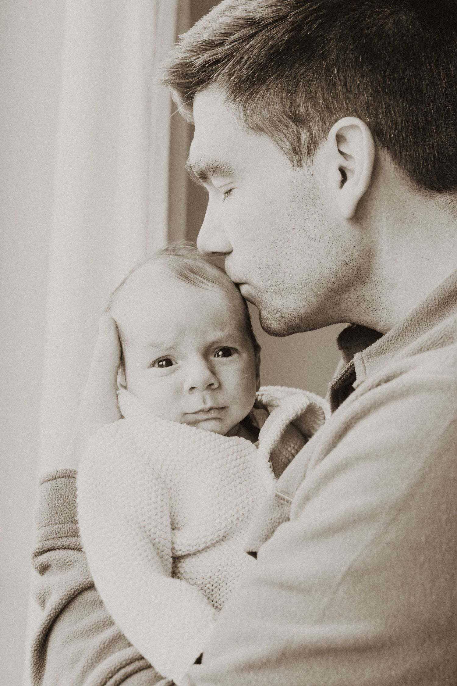 Black and white photo of a father tenderly kissing his newborn baby on the head while cradling them gently.