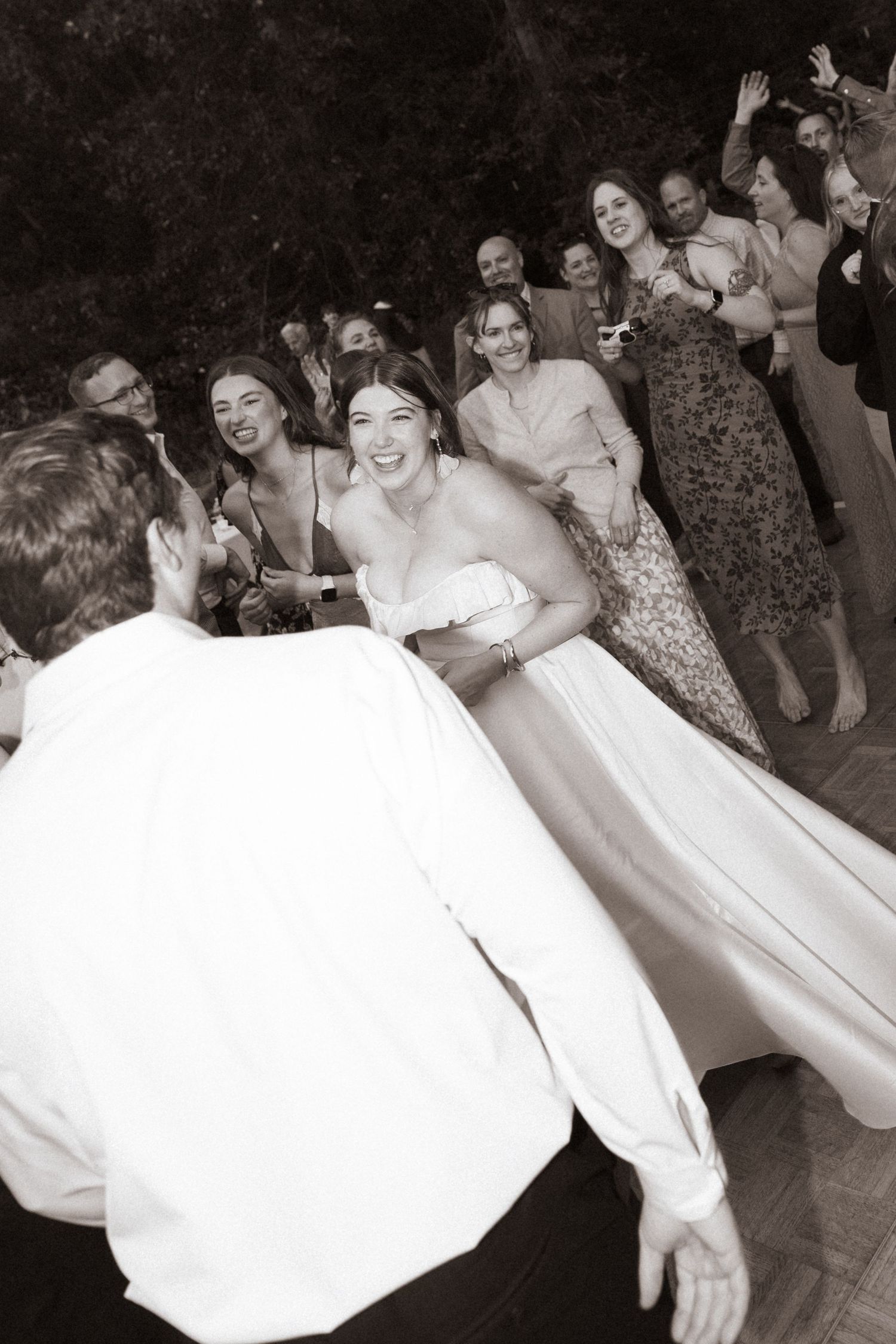 Joyful bride laughs with wedding guests during lively outdoor reception dance.
