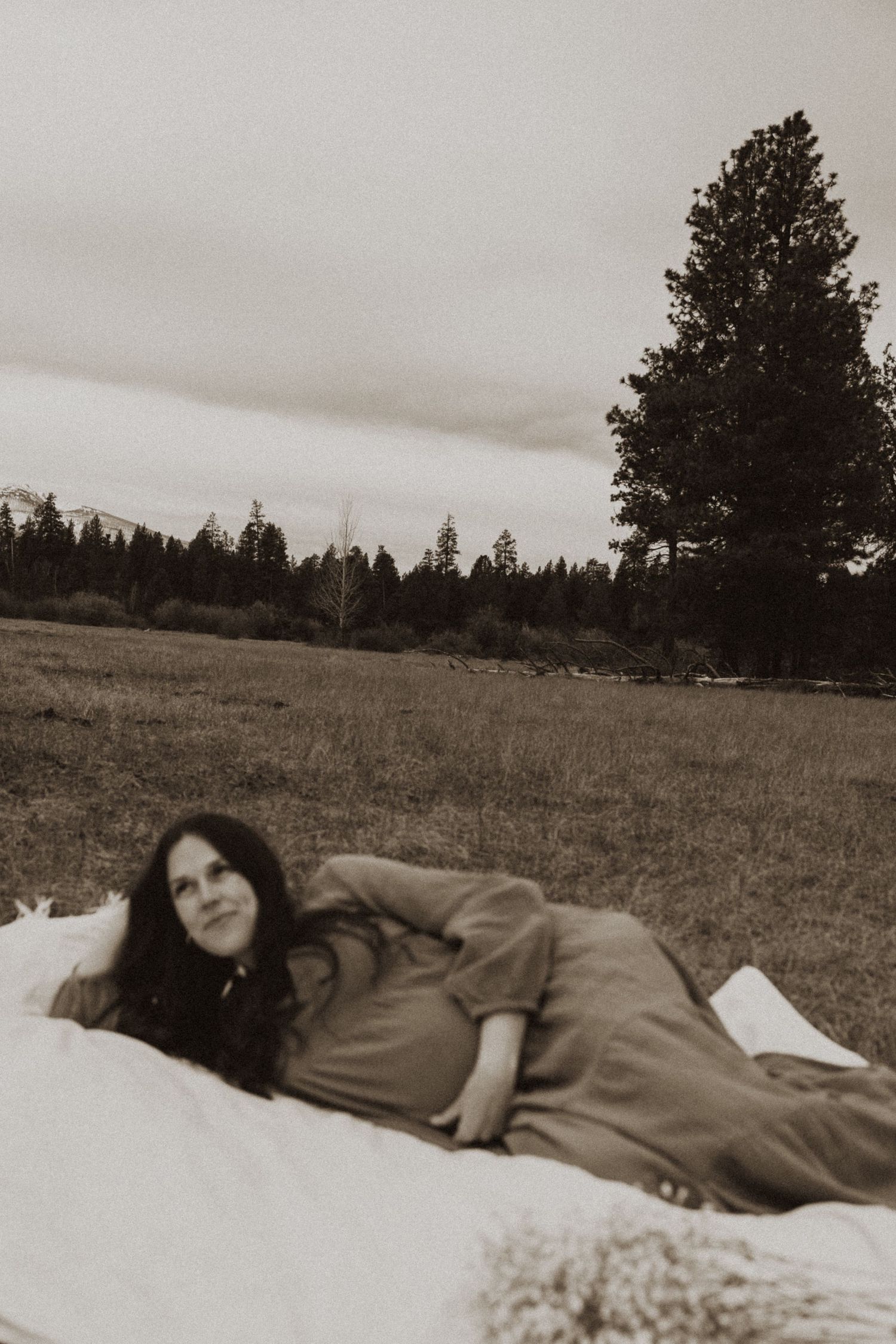 Pregnant woman in olive dress lying on white blanket in open field with pine trees under overcast sky.