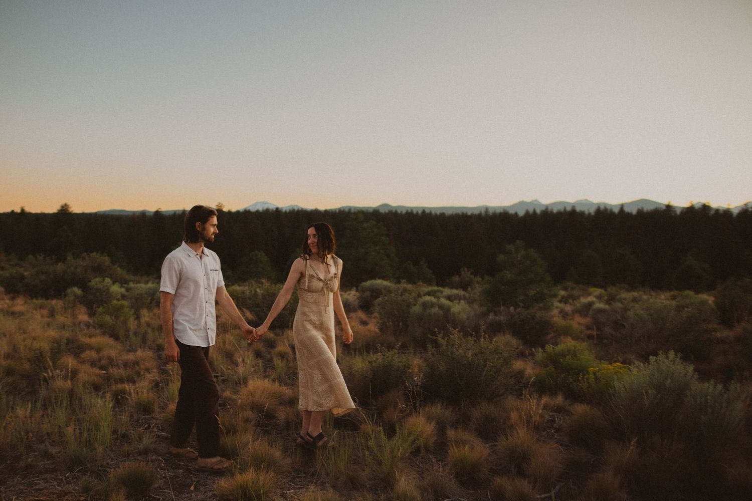 A couple walks hand in hand through desert brush during sunset.
