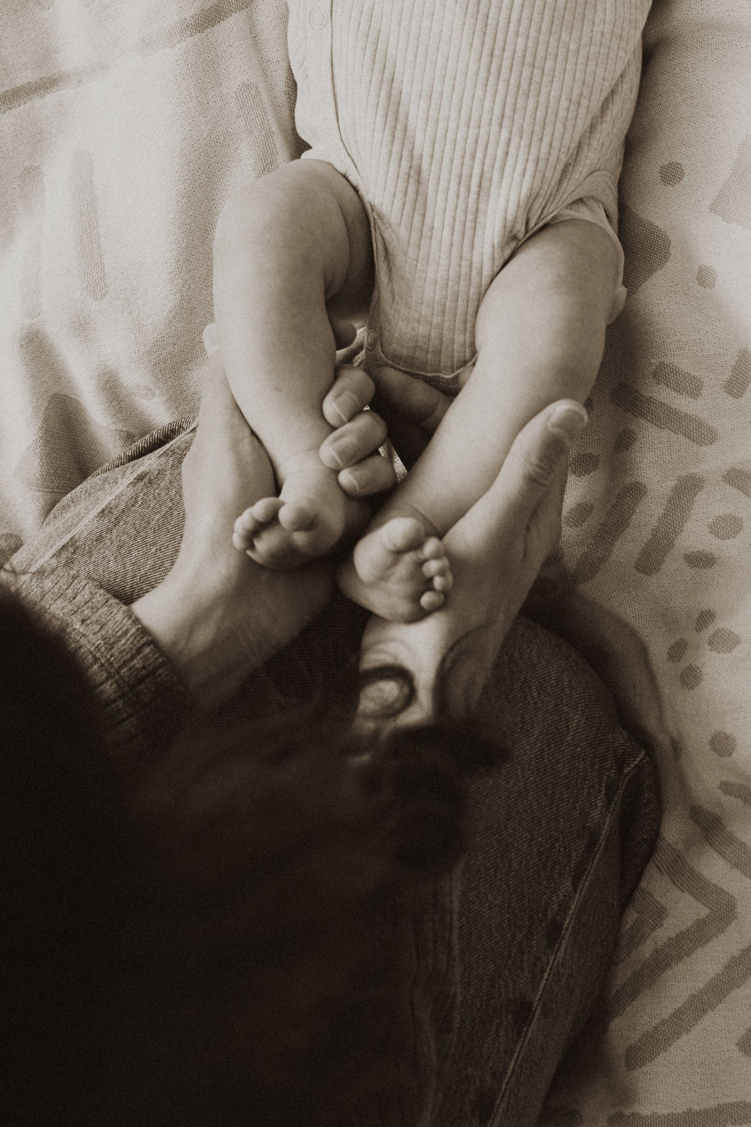 Sepia-toned close-up of a parent gently holding a newborn baby's tiny feet and toes on a soft blanket.