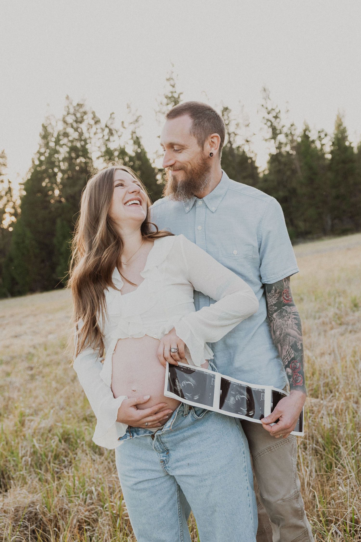 Smiling pregnant couple holding ultrasound photos in a sunny meadow, man with tattoos embracing woman touching her baby bump.