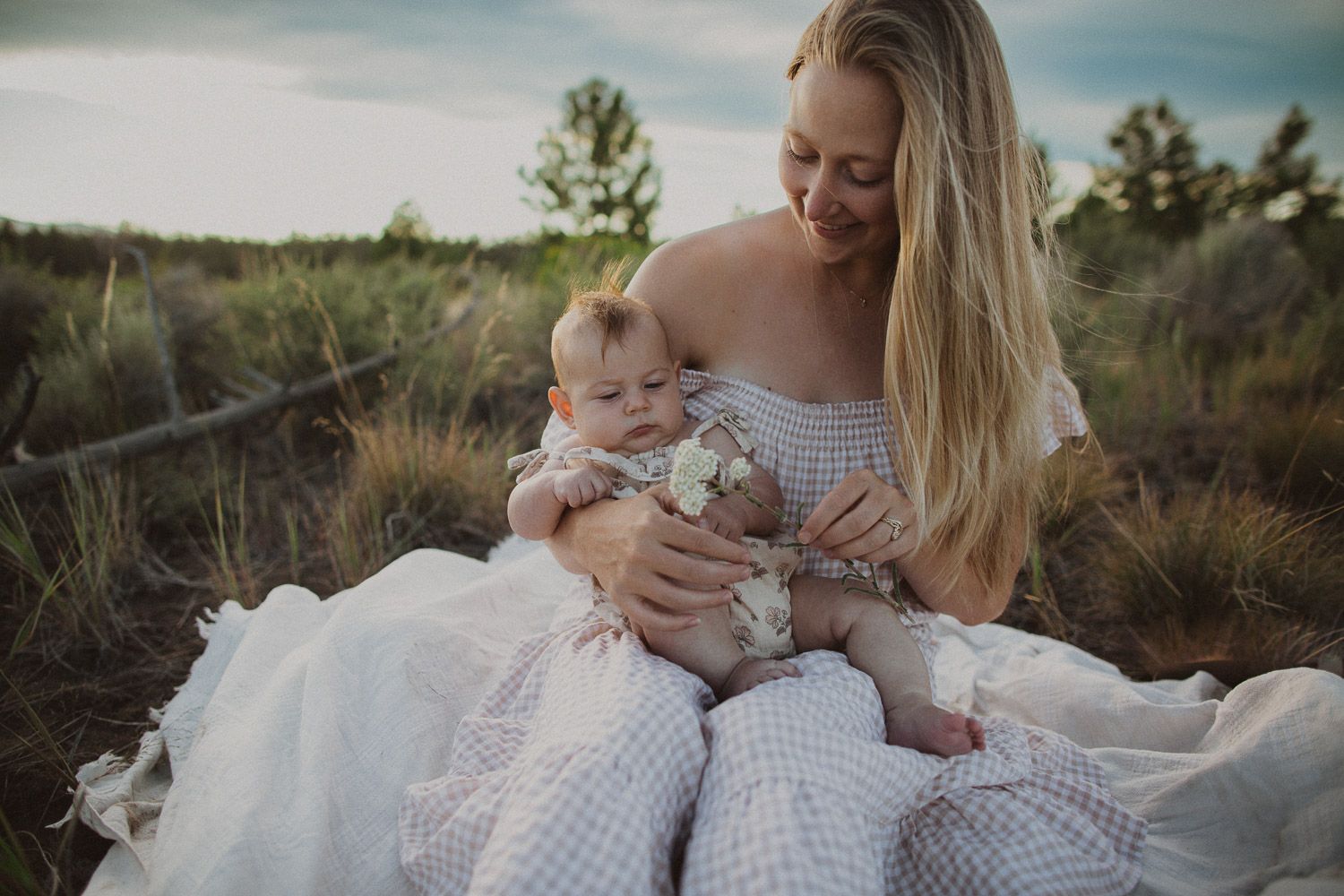 A tender outdoor portrait of parent and infant sitting on a white blanket in a natural field setting at dusk.