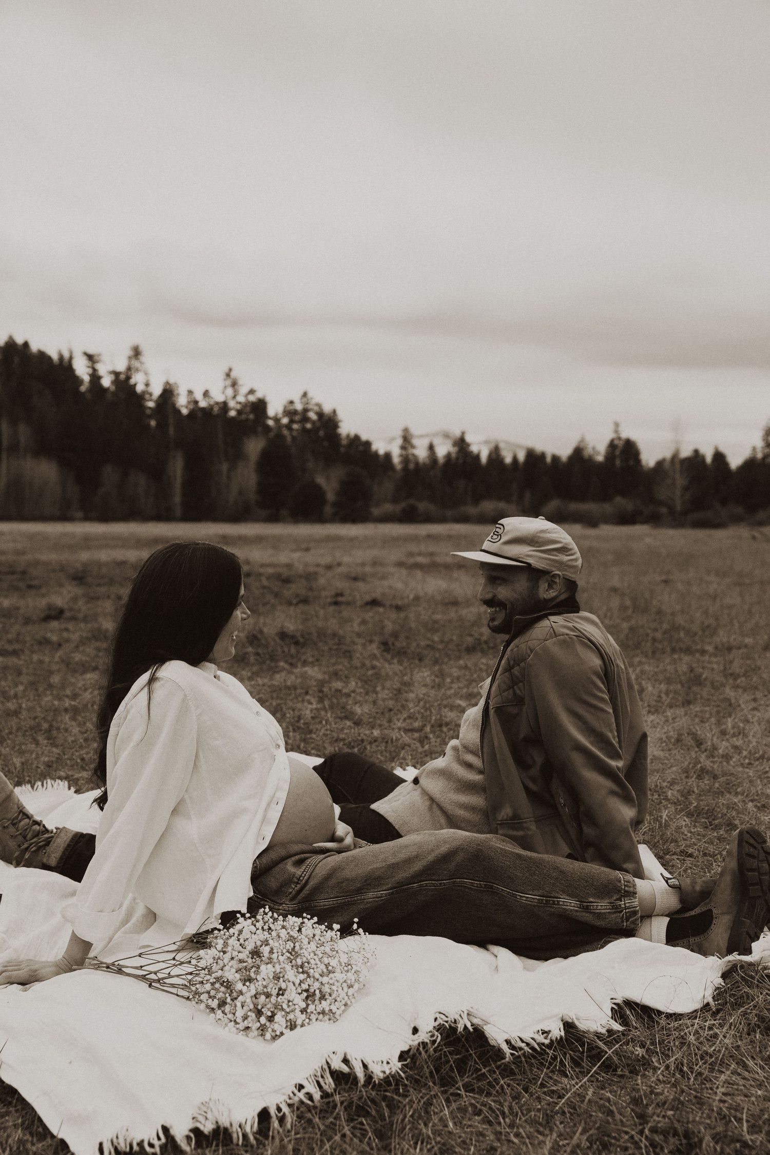 Black and white maternity photo of couple sitting on blanket in open field with baby's breath bouquet.