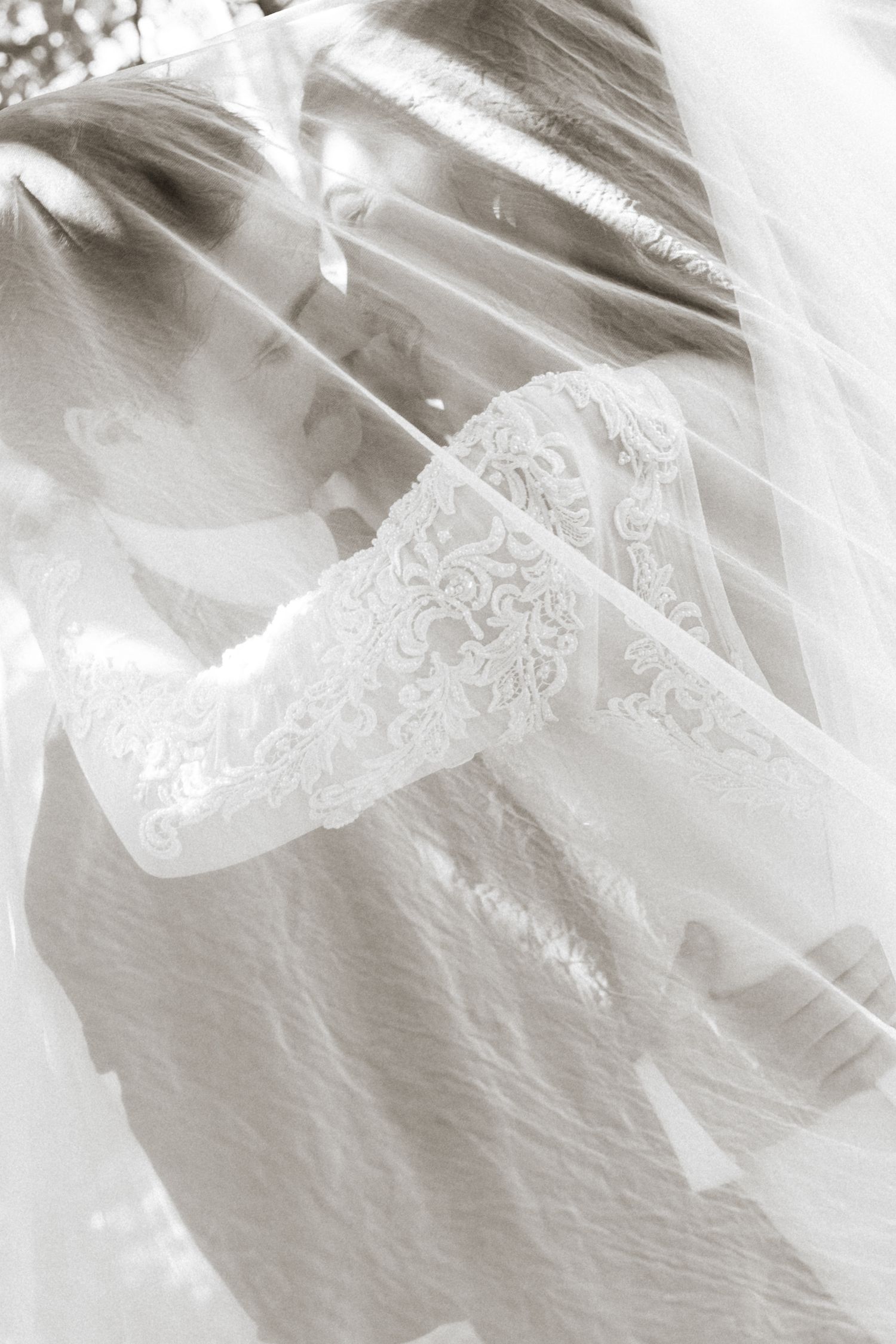 Bride and groom share an intimate kiss beneath a flowing white wedding veil in a soft sepia-toned portrait.