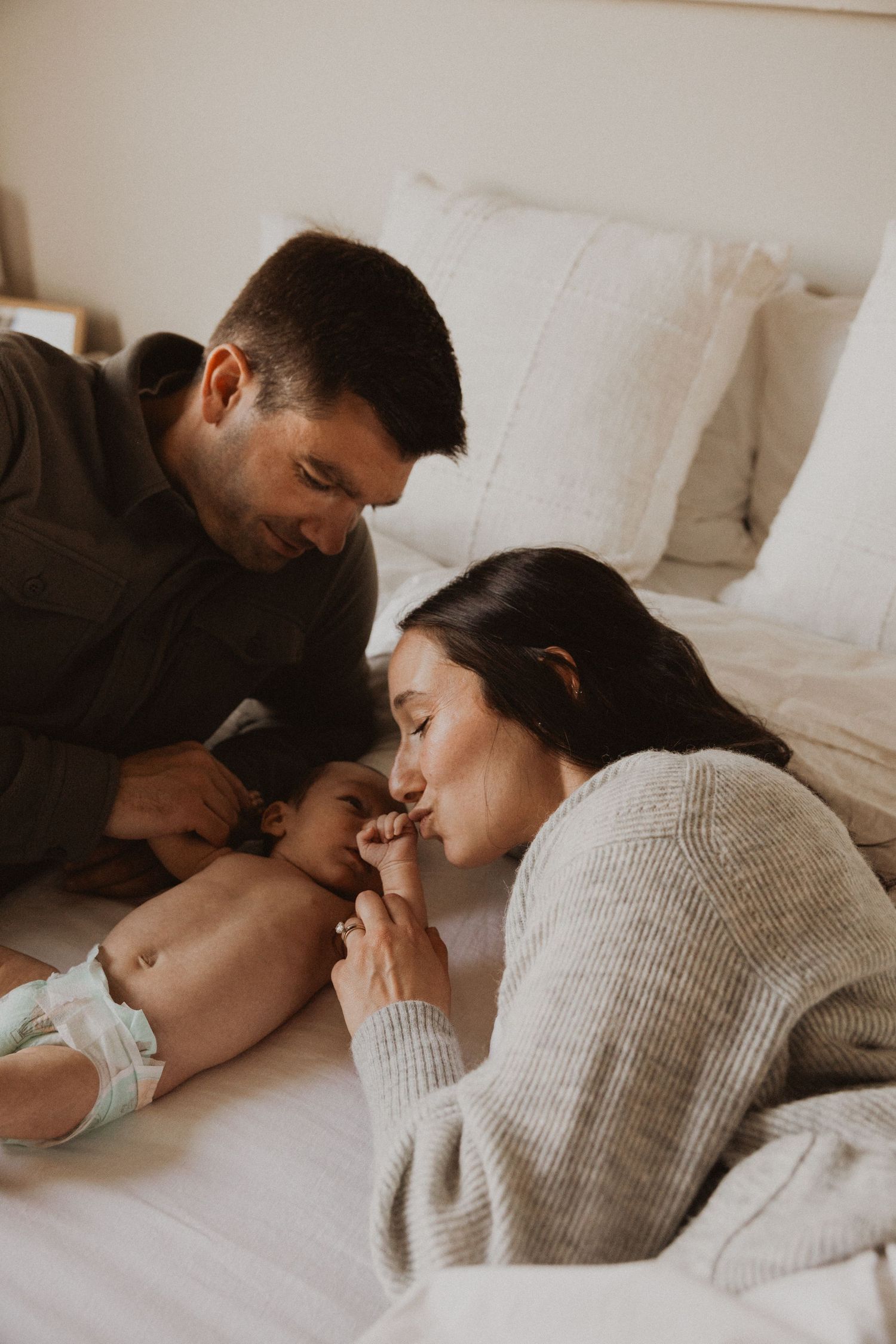 Parents lovingly tend to their newborn baby on a cozy white bed in warm natural light.