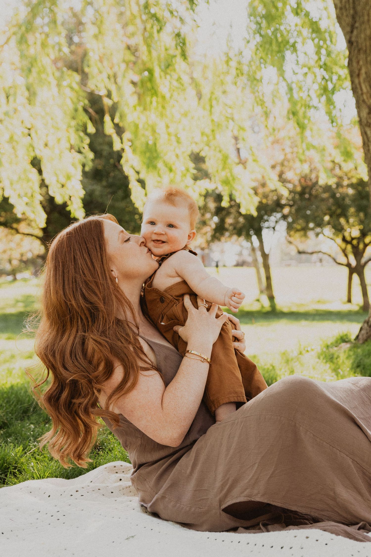 A mother kisses her smiling red-haired baby outdoors on a blanket under a willow tree in warm sunlight.