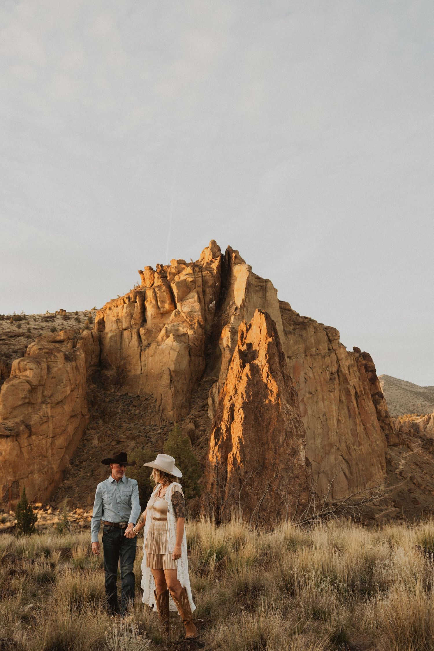 Couple holding hands walking through desert landscape with dramatic red rock formations at golden hour.