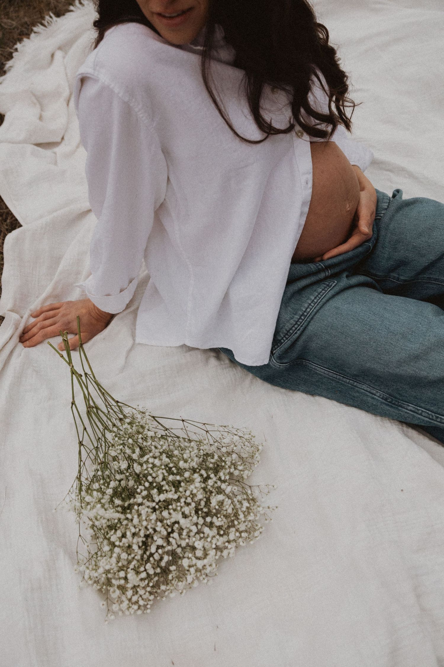 Overhead view of pregnant woman in white shirt sitting on blanket holding a baby's breath bouquet.