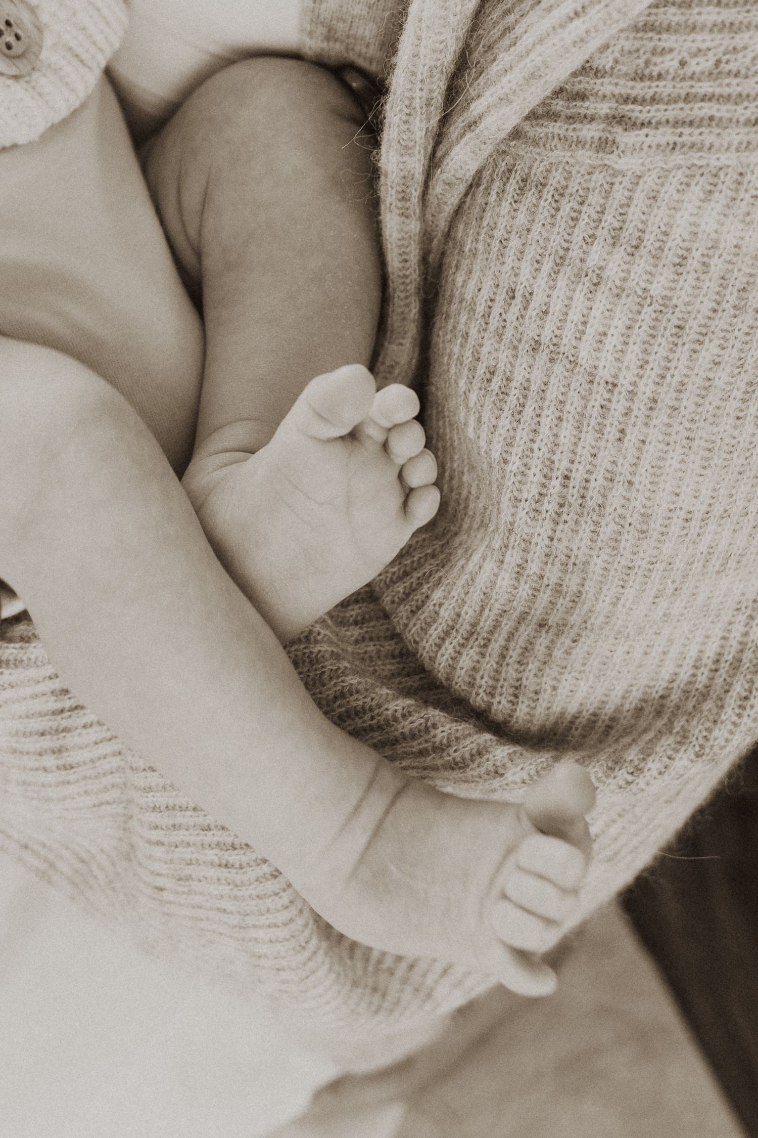 Sepia-toned close-up of tiny newborn baby feet resting against a soft knit sweater held by a parent.