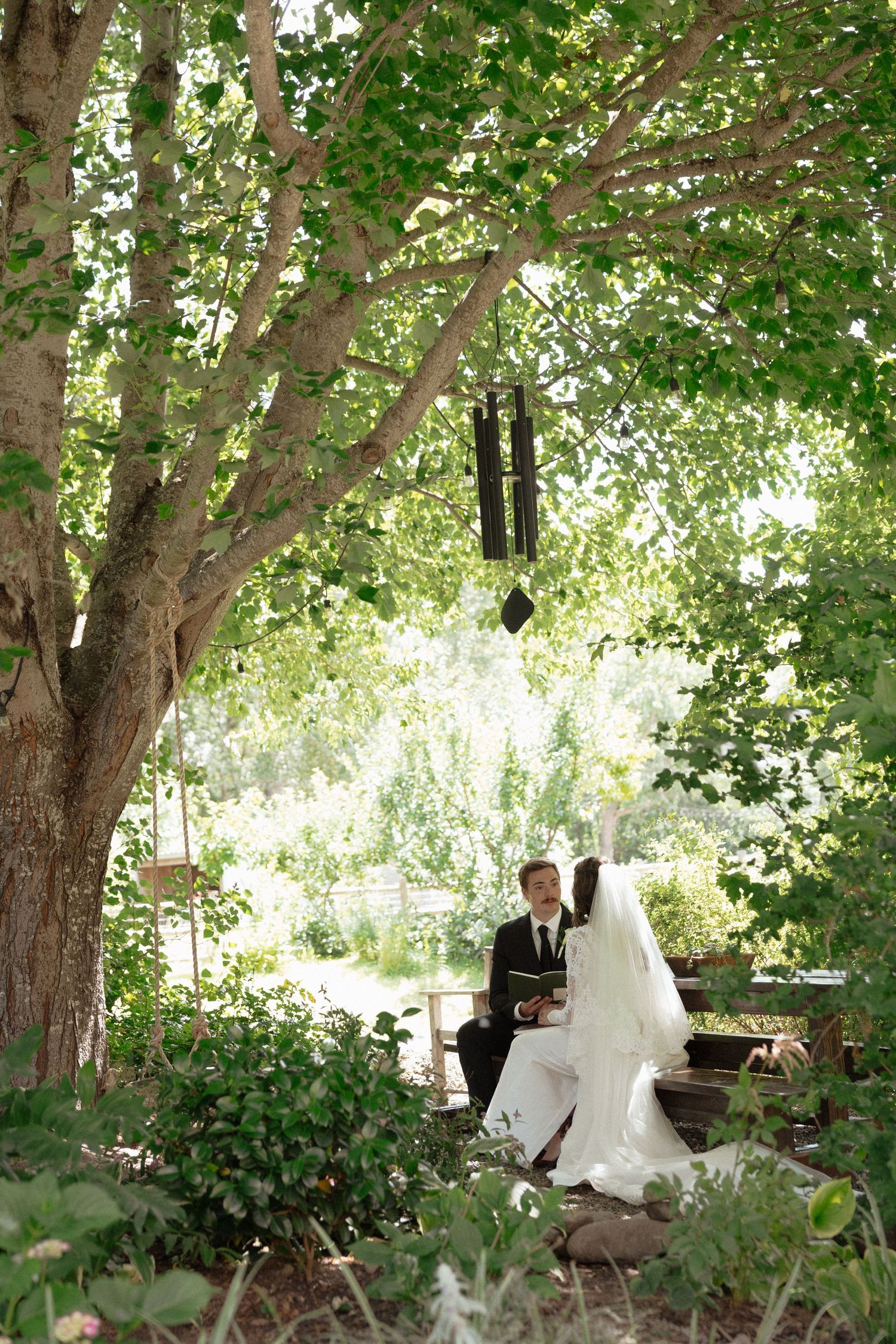 Bride and groom seated under a large leafy tree during an intimate outdoor garden wedding ceremony.