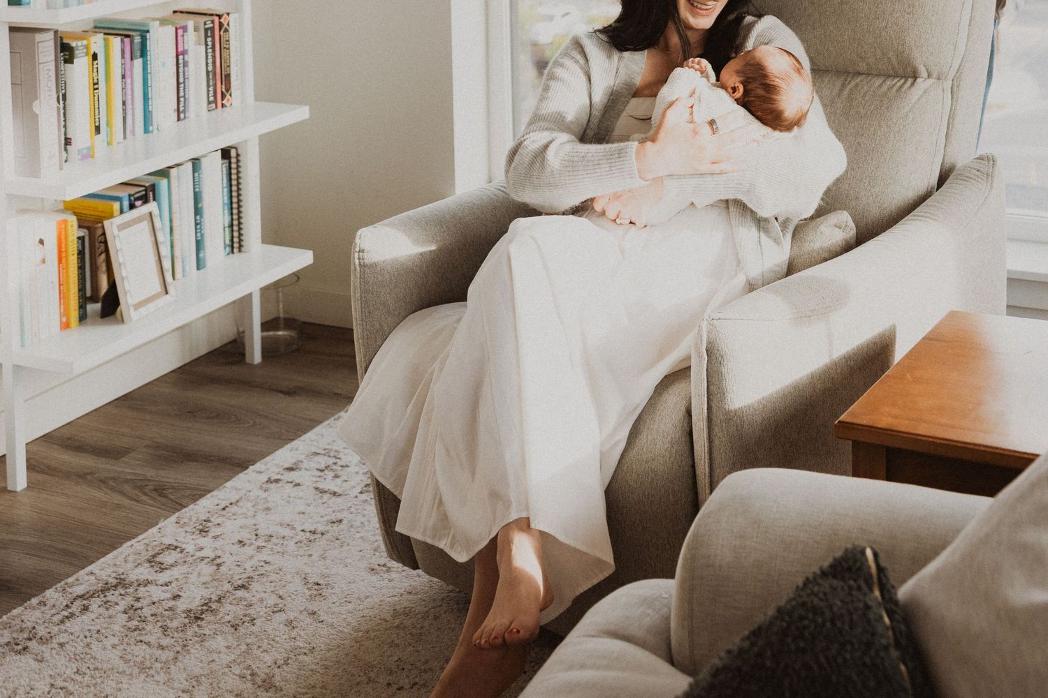 Mother sitting in gray armchair holding newborn baby in bright living room with bookshelf.