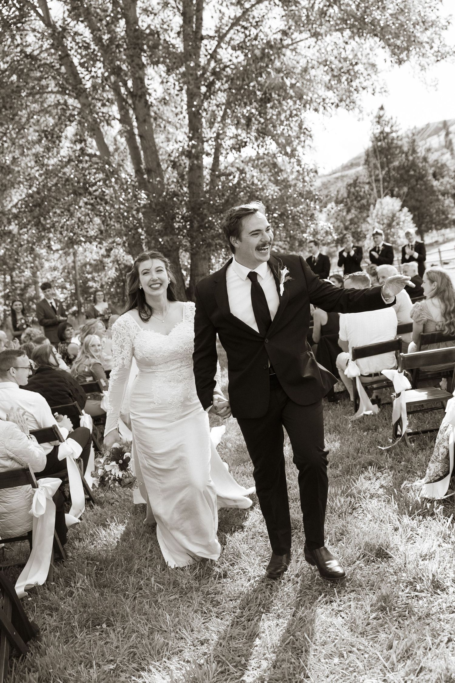 Joyful newlyweds walk hand-in-hand down the aisle after their outdoor wedding ceremony in black and white photo.