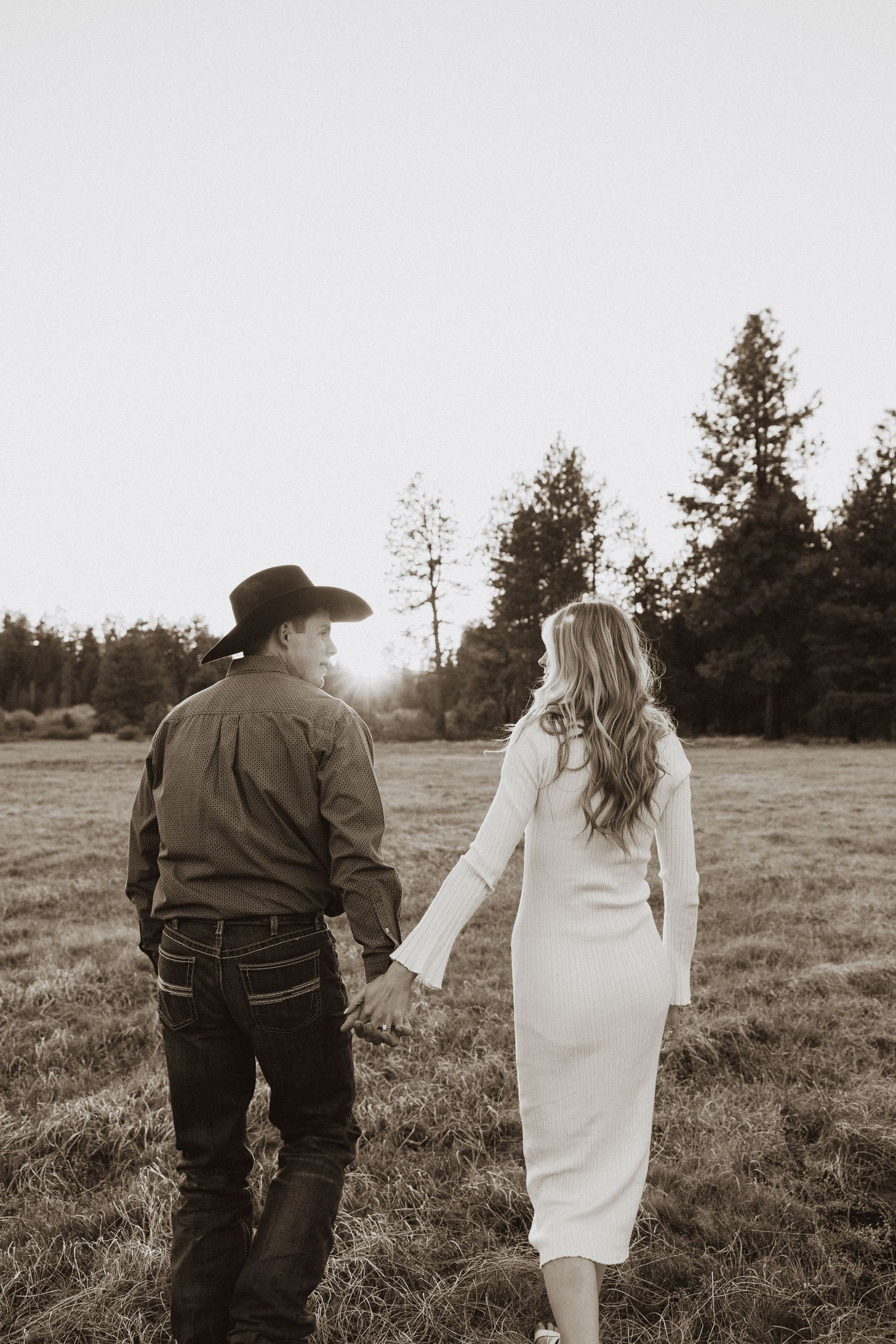 Couple holding hands walking through a field at sunset, man in cowboy hat and woman in white dress, sepia tone.