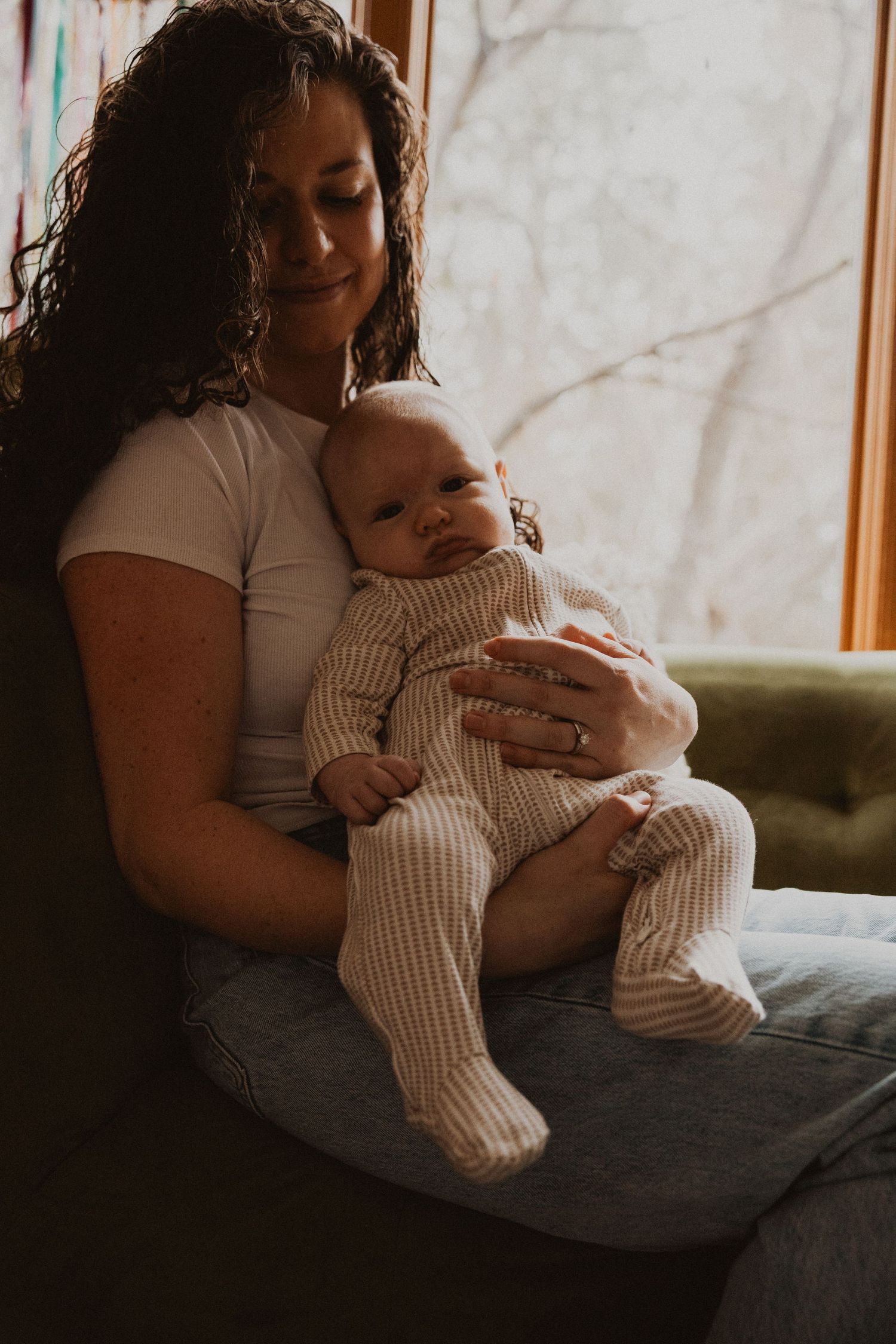 Curly-haired mother smiling down at newborn baby in striped onesie on green couch.