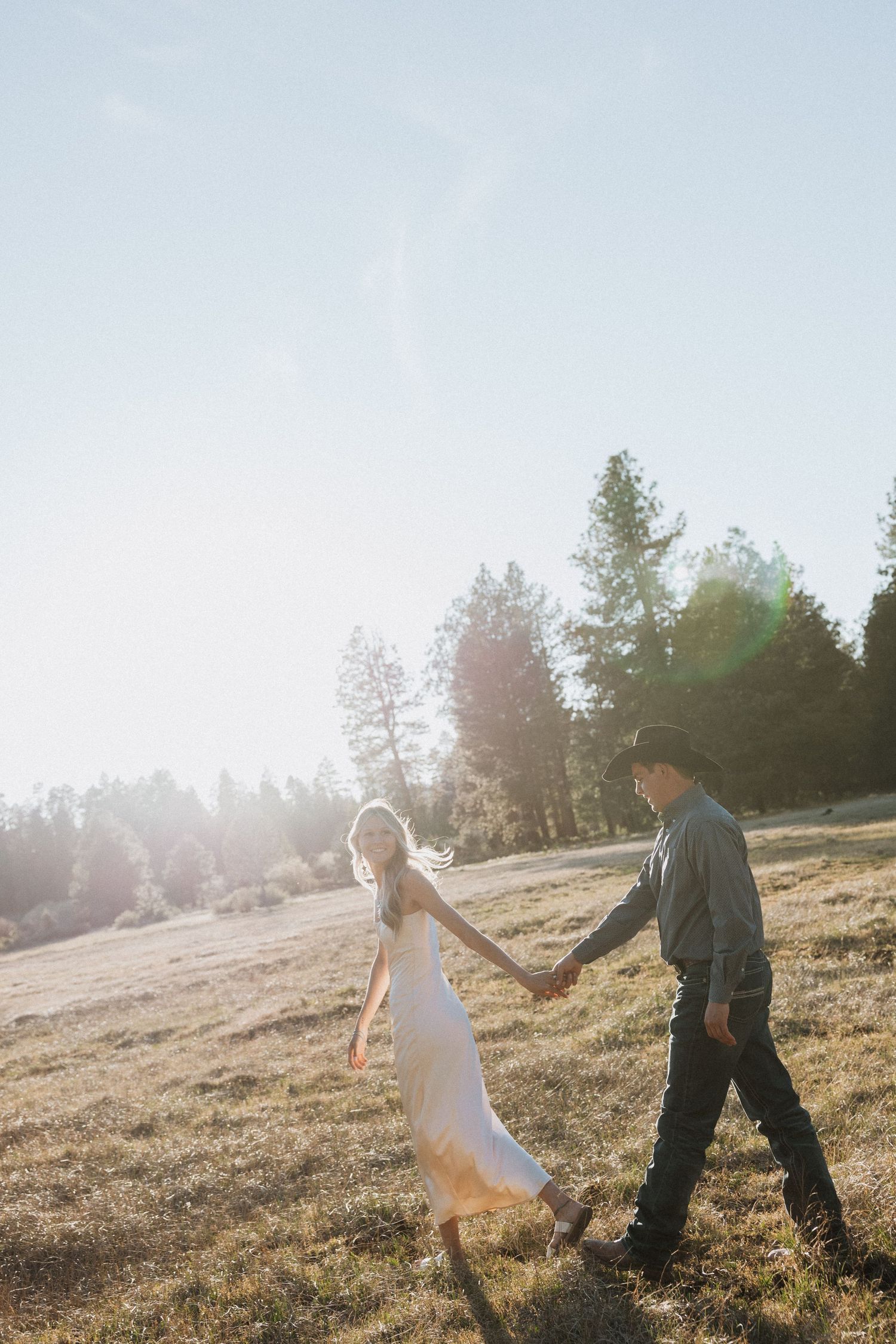 Couple holding hands walking through a sunlit meadow with pine trees, woman in white dress, golden hour light.