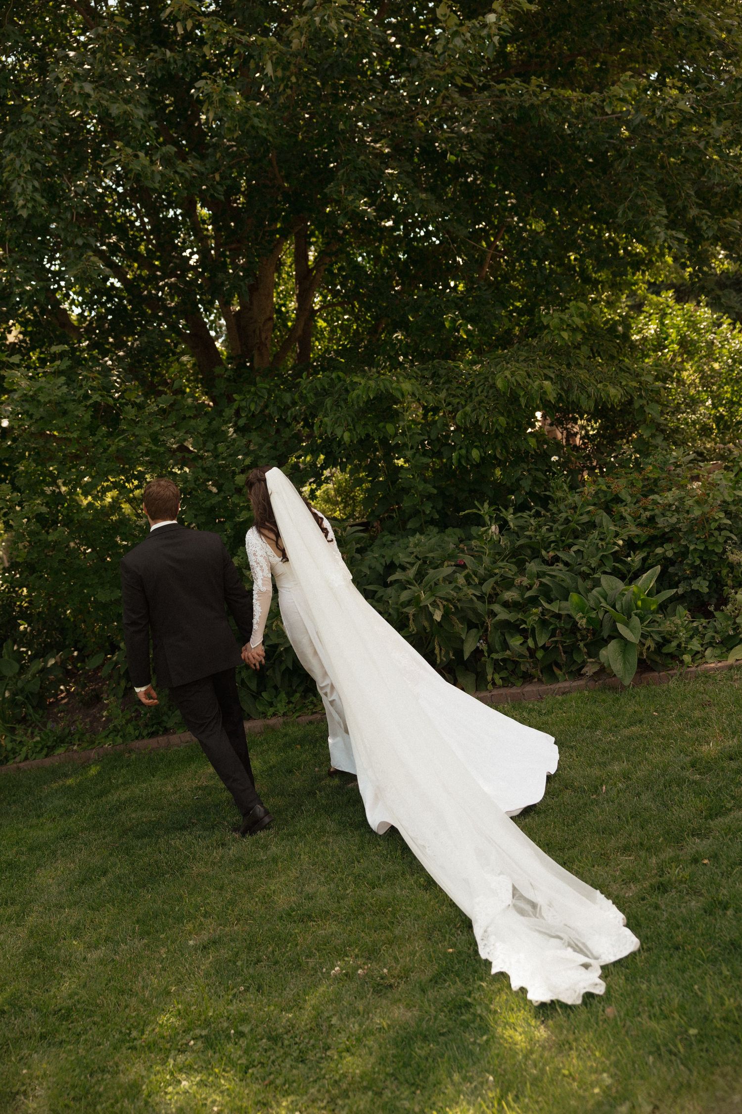 Bride and groom walking hand in hand through lush green garden, wedding veil and train flowing behind.