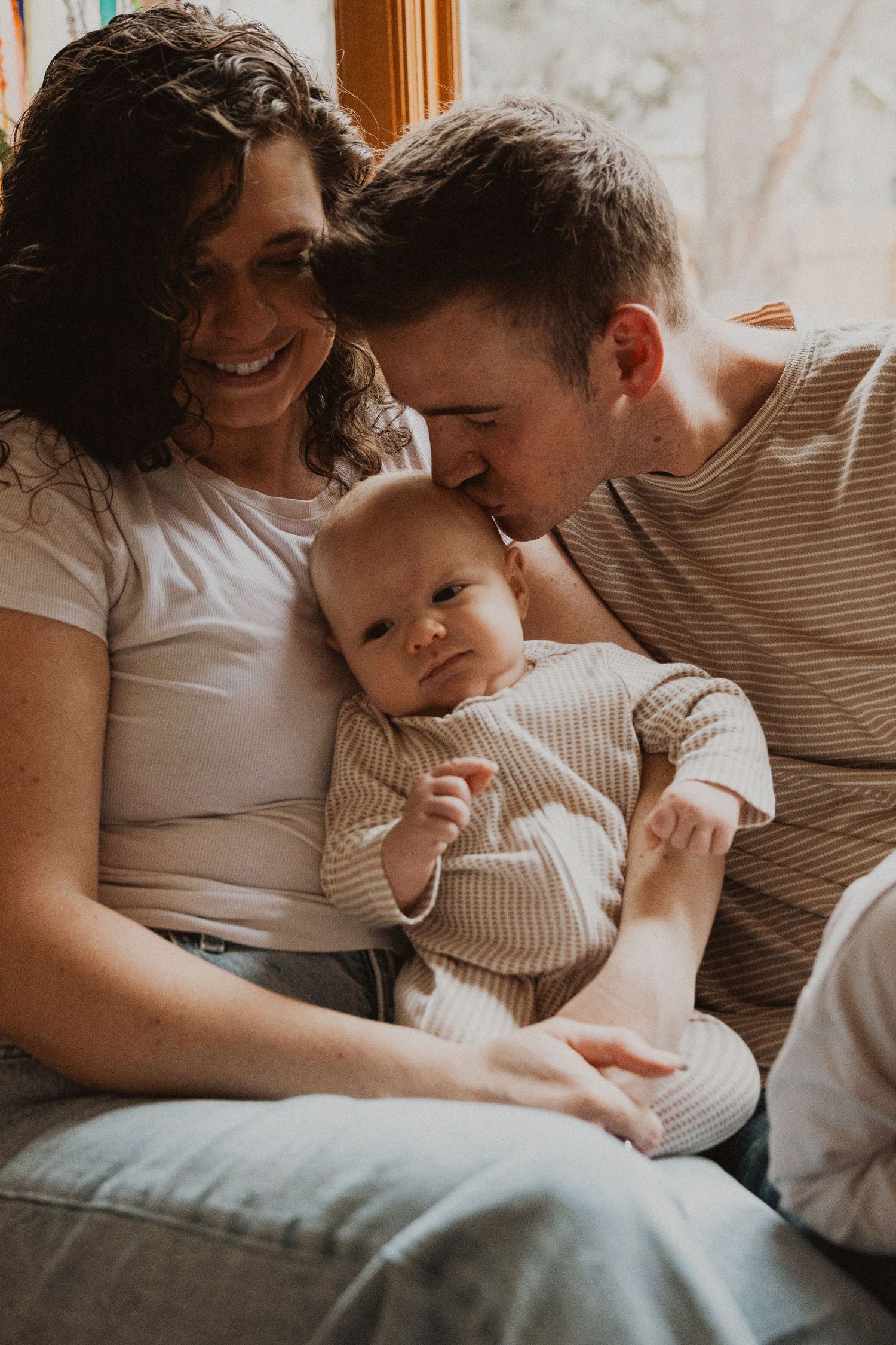 Happy parents kissing and cuddling their newborn baby together on a cozy couch at home.