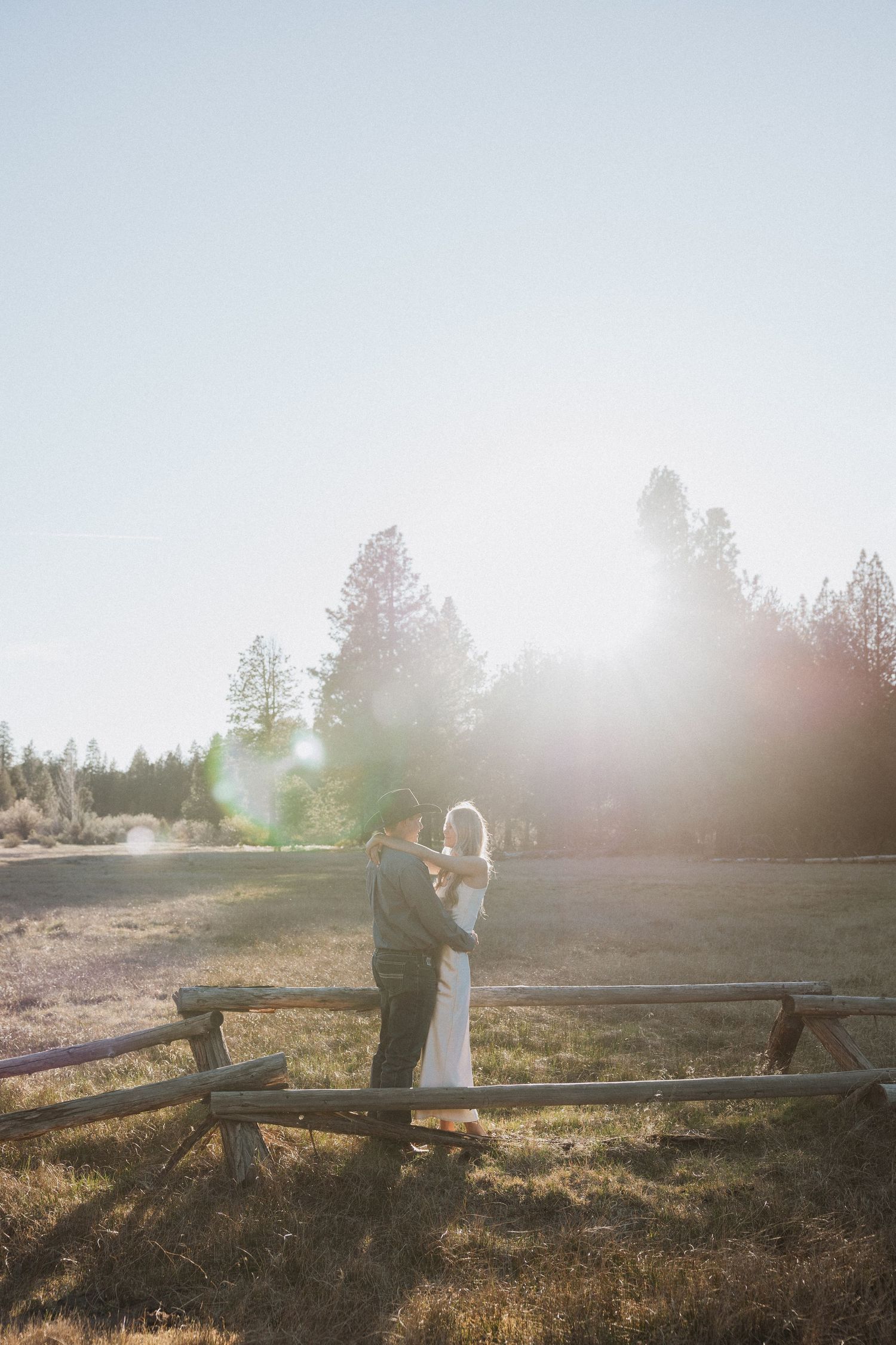 Bride and groom embrace in golden sunlight by a rustic wooden fence with tall pine trees in background.
