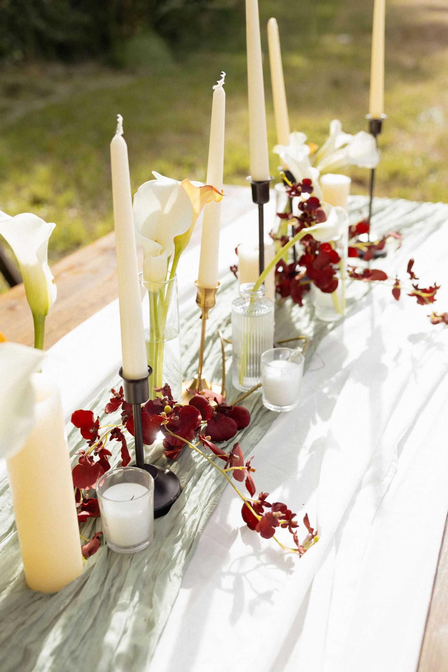 Elegant wedding reception table adorned with white candles, calla lilies, and deep red orchids as centerpieces outdoors.