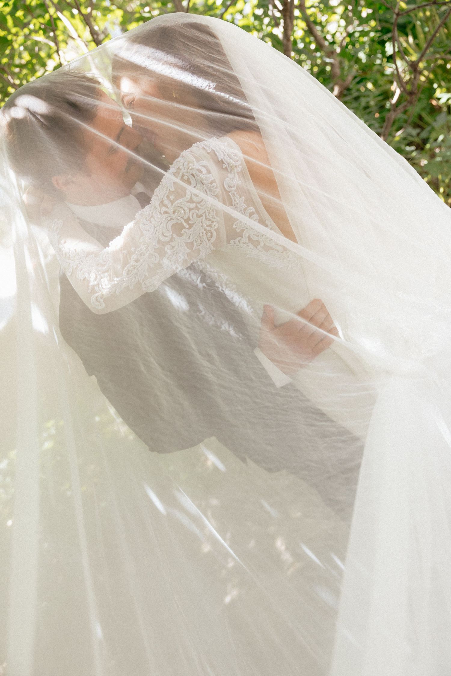 Bride and groom sharing a romantic kiss under a flowing white wedding veil outdoors surrounded by lush green trees.