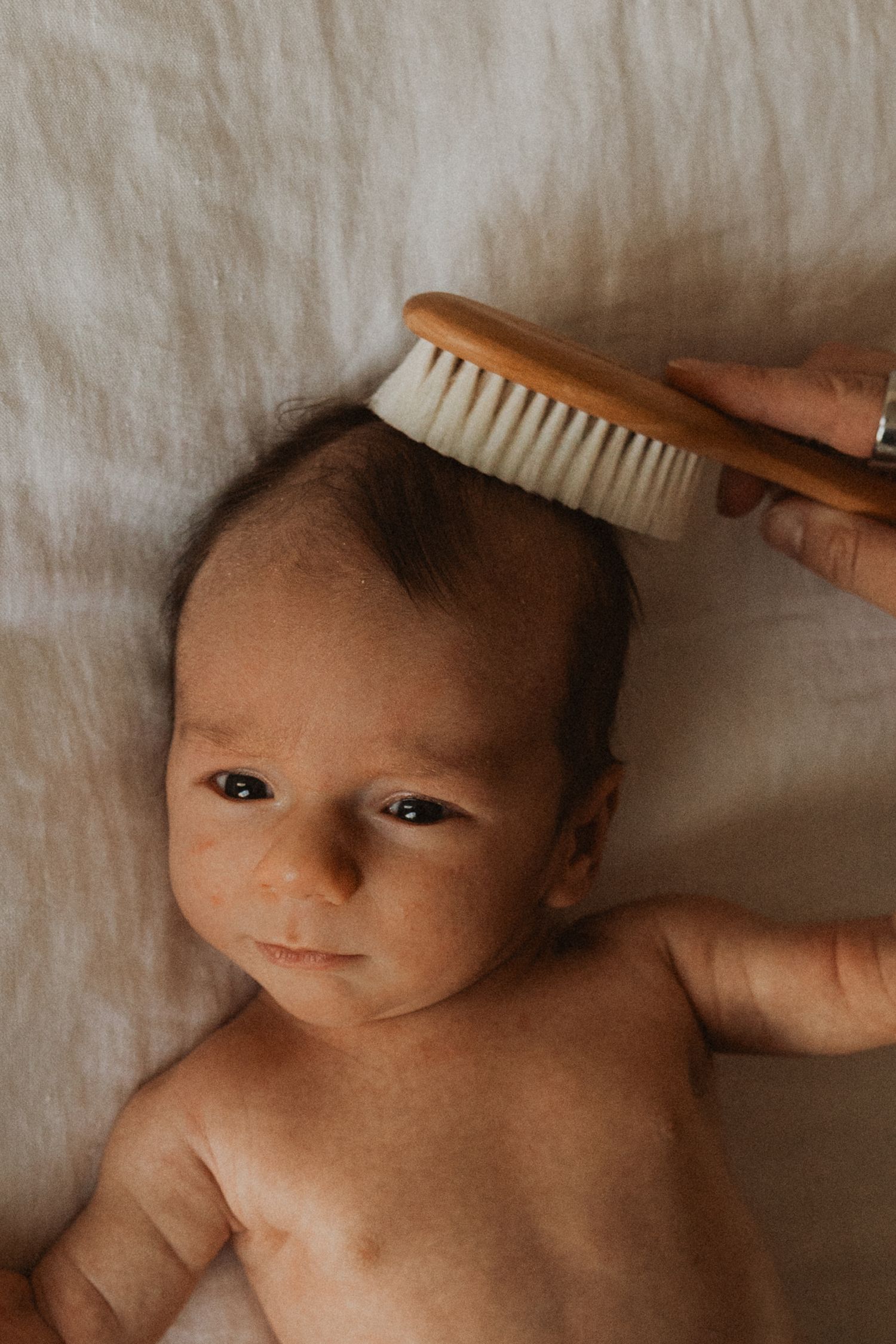 A newborn baby lying on a white blanket while a hand gently brushes their dark hair with a wooden bristle brush.