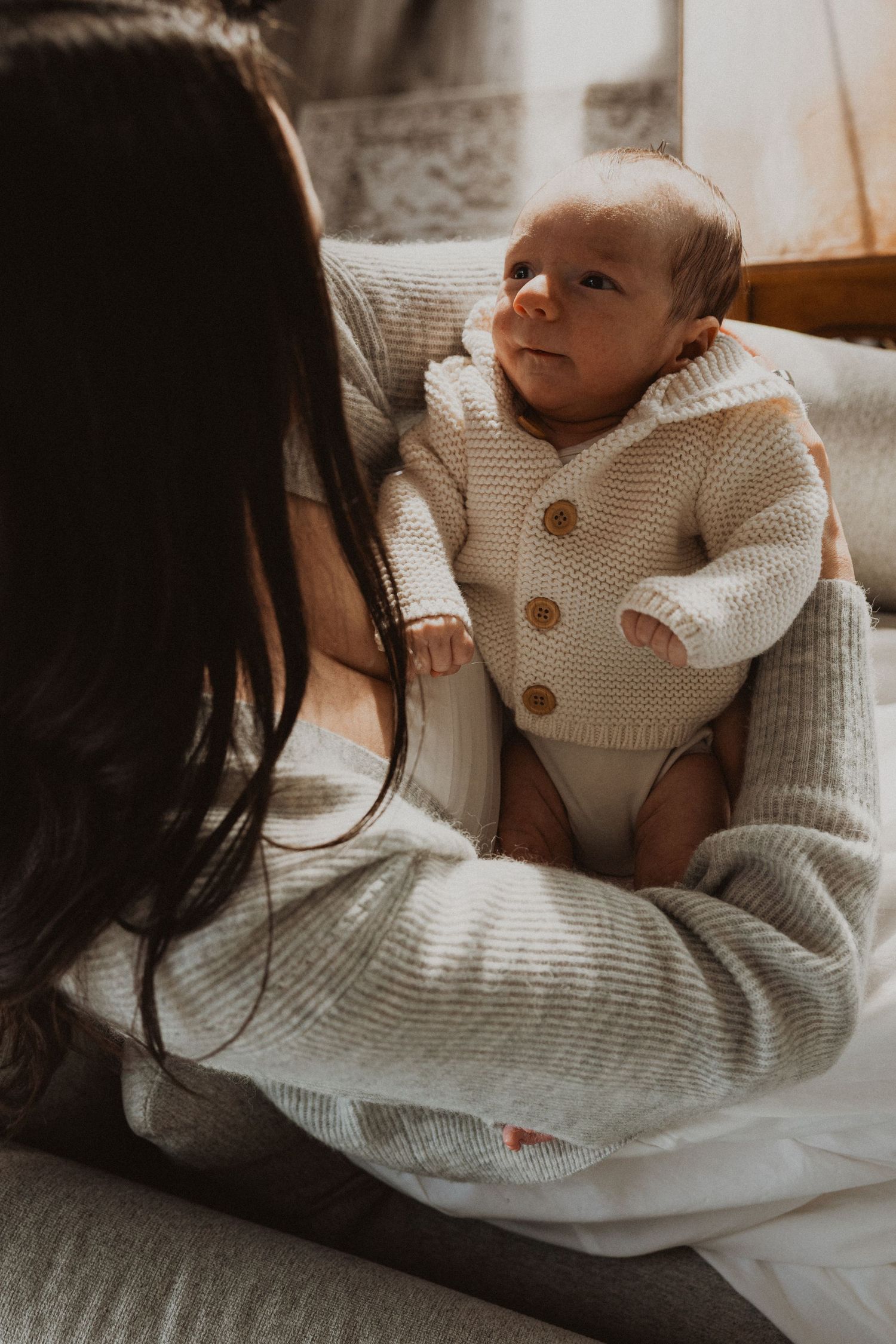 A mother in a knit sweater holds her newborn baby dressed in a cream knit cardigan in a warmly lit room.