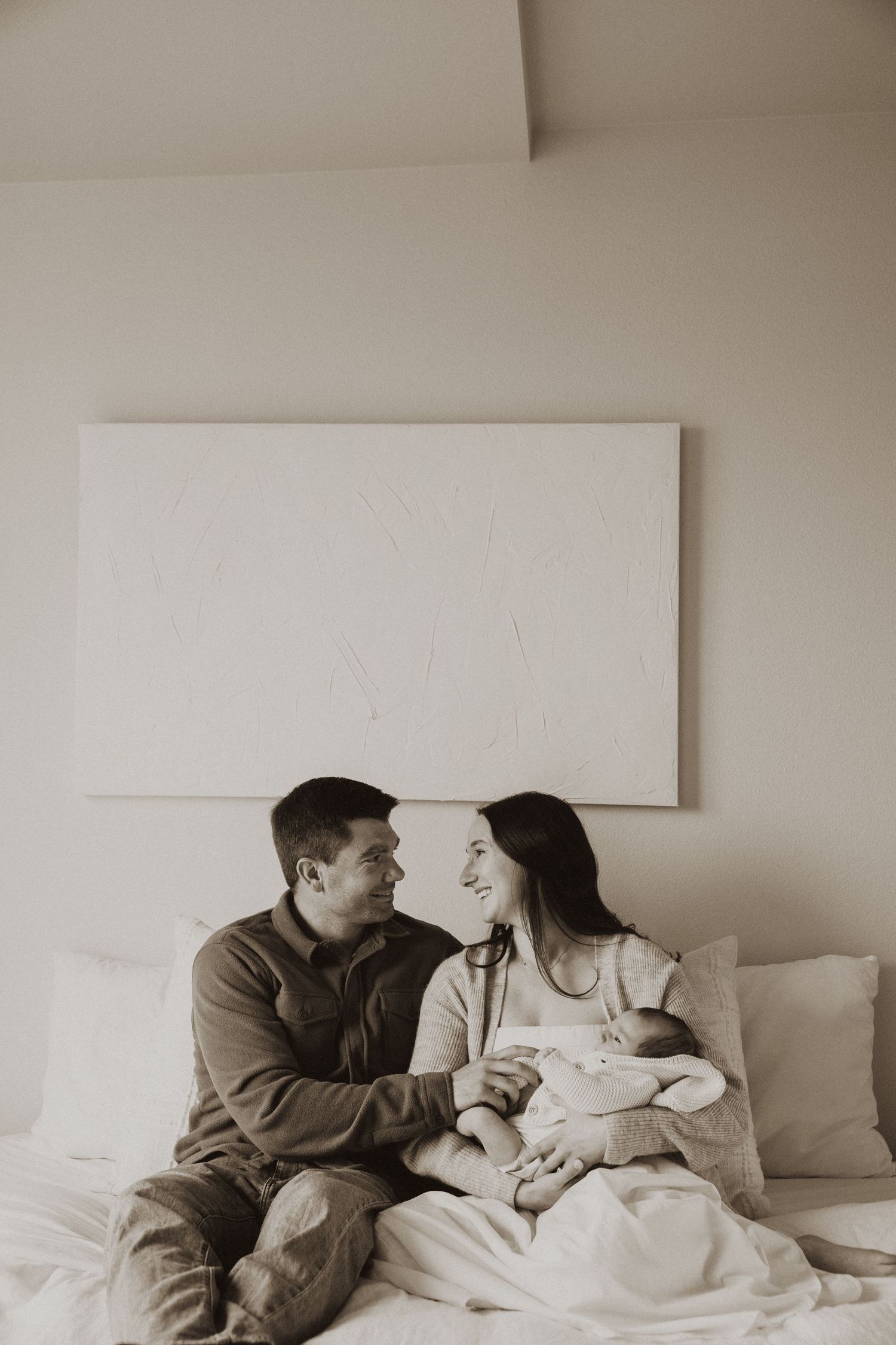 A couple sits on a bed smiling at each other while cradling their newborn baby in a serene black and white photo.