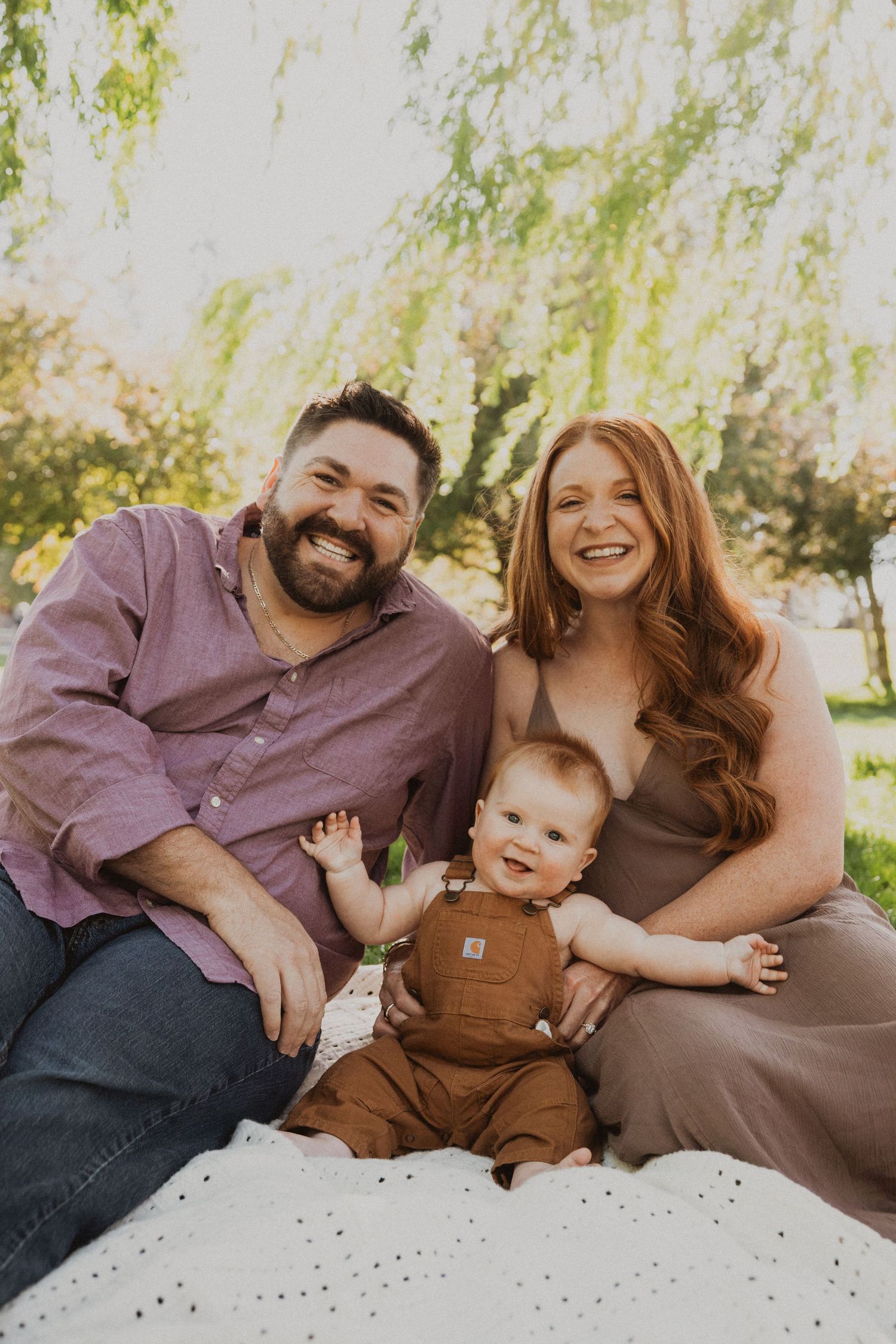 A smiling family with a baby in brown overalls sits on a blanket under a willow tree outdoors.