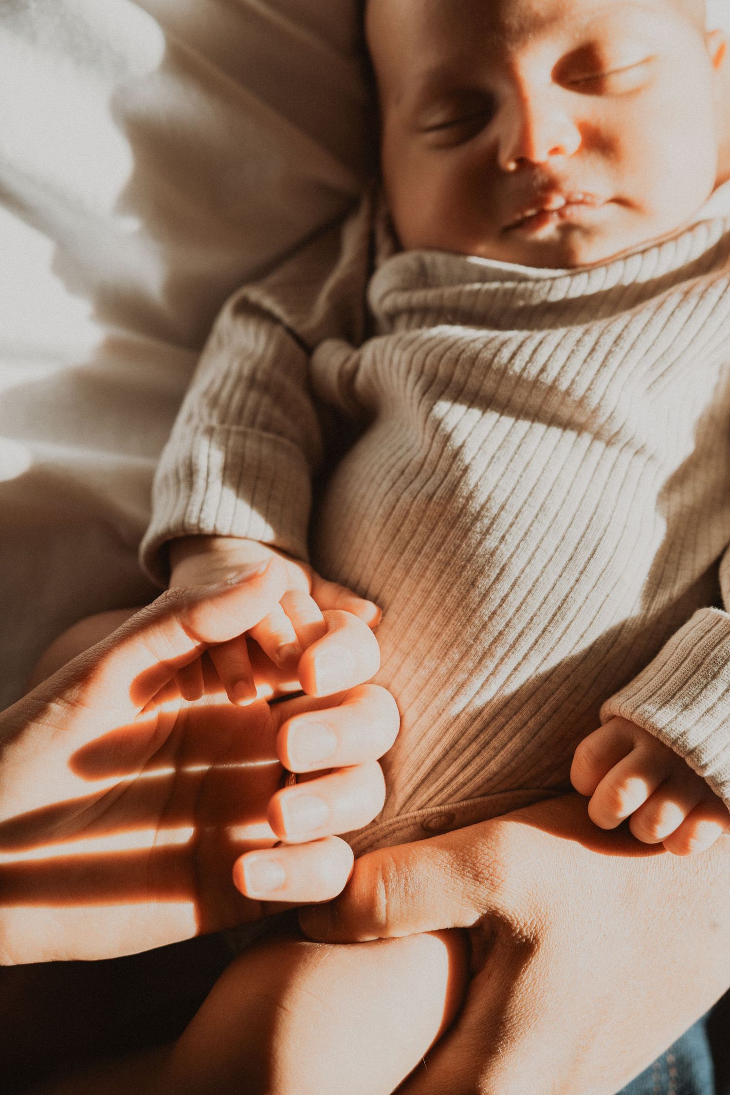 Newborn baby sleeping peacefully in warm sunlight, tiny hand gently held by parent in soft ribbed onesie.