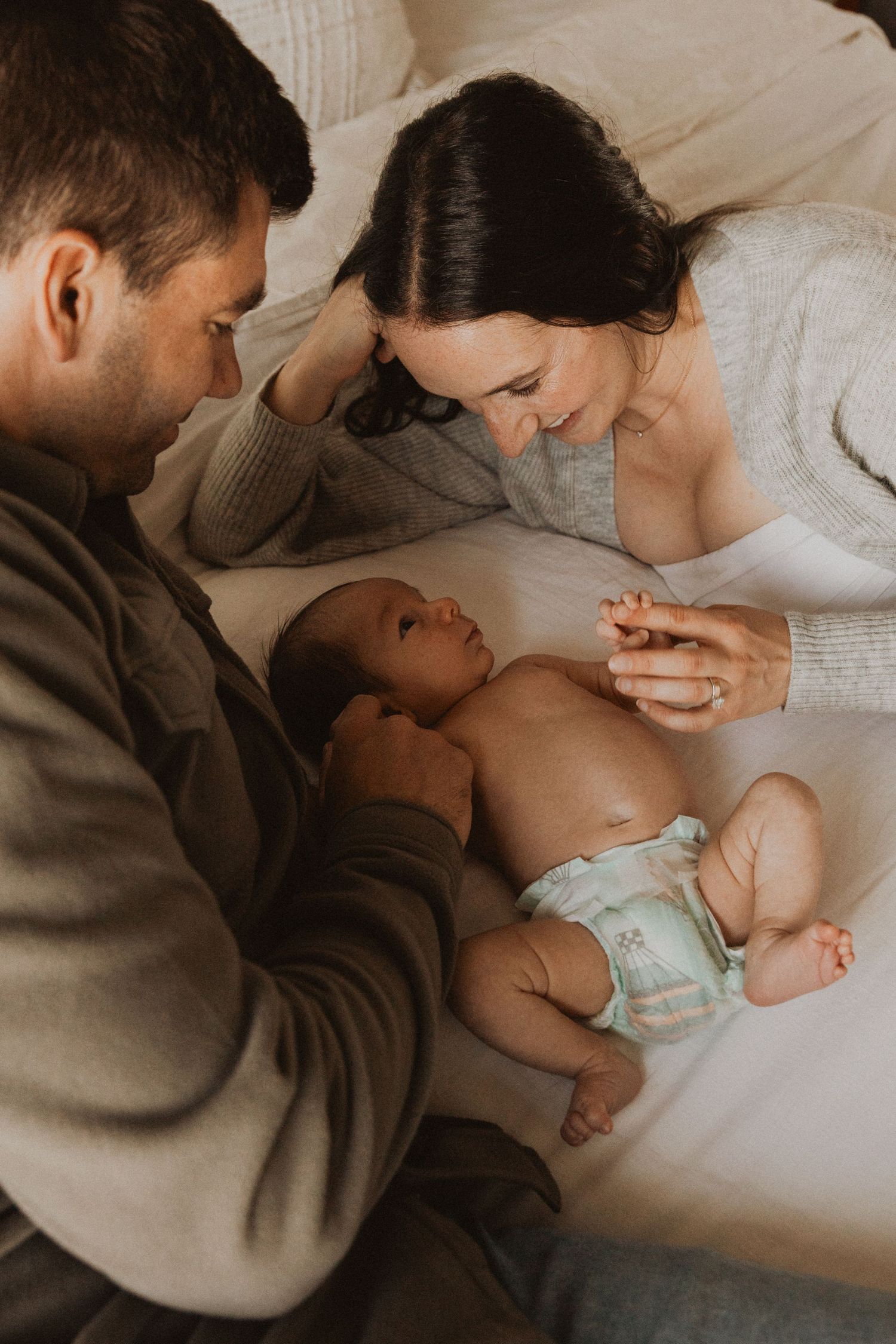 Parents lovingly gazing at their newborn baby lying on a white bed, in a warm intimate family portrait.