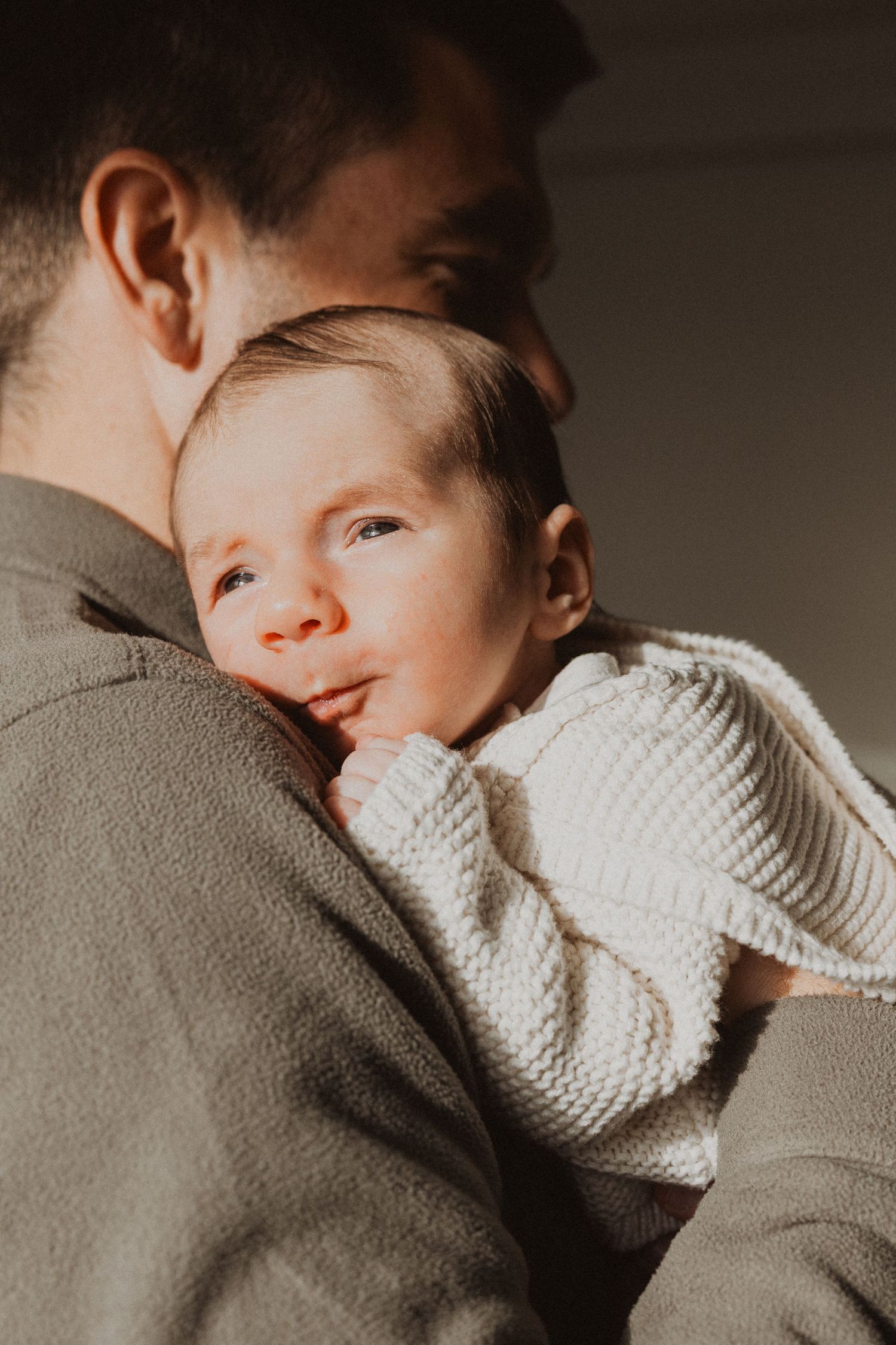 A tender moment of a father cradling his newborn baby dressed in a white knit sweater in warm golden light.