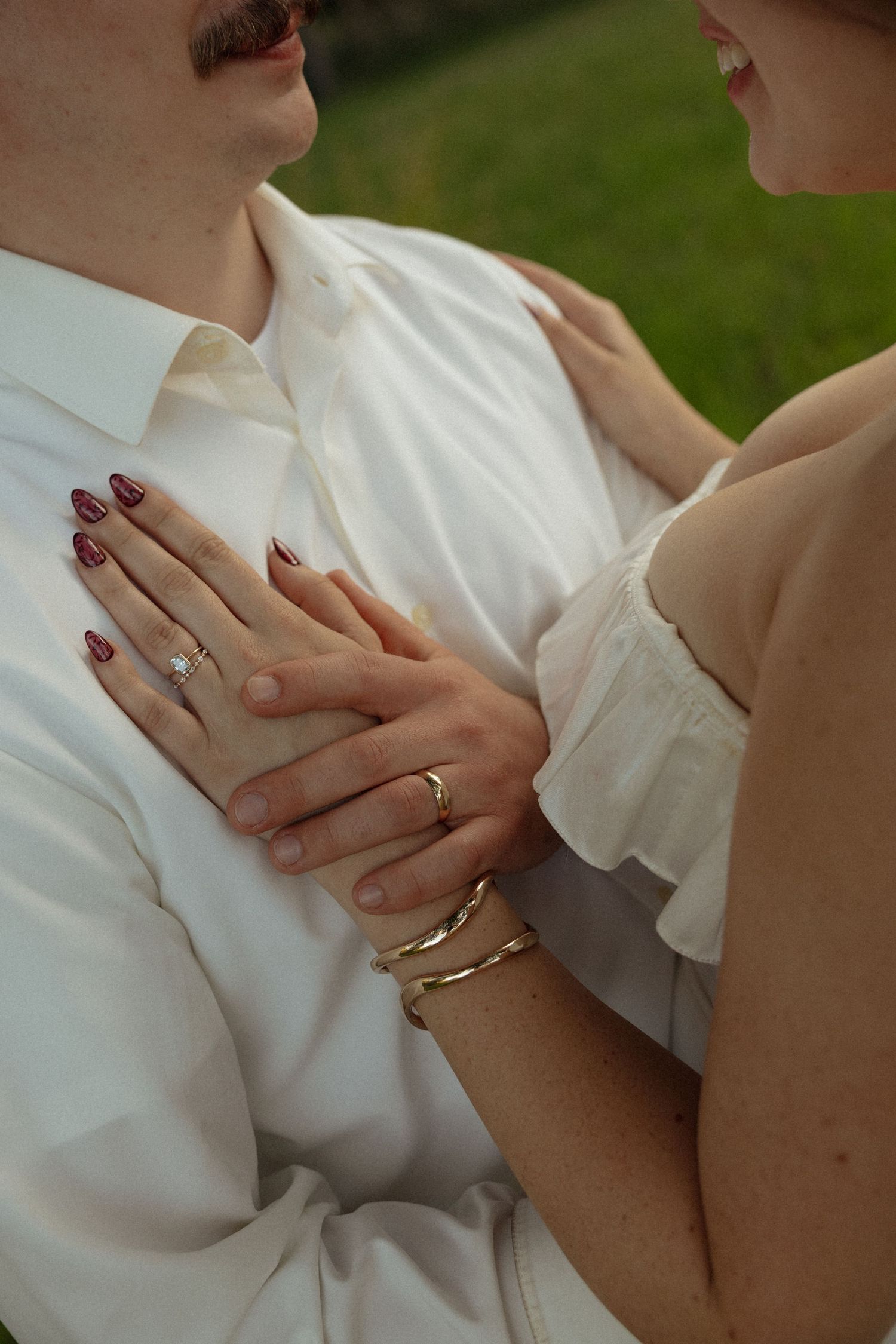 Close-up of bride's hands with diamond ring and gold bracelet resting on groom's white shirt chest.
