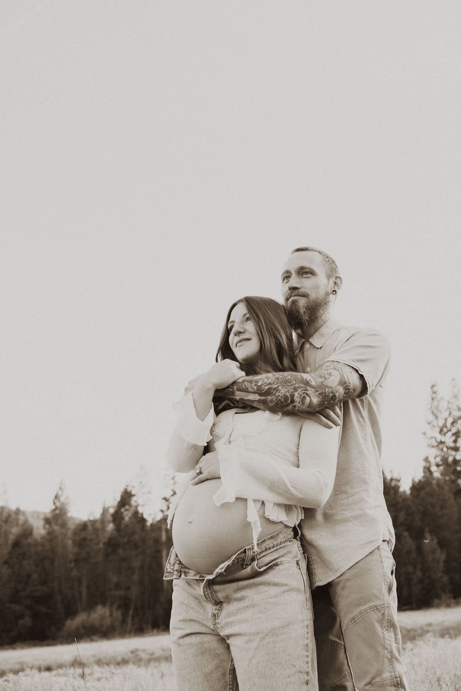 Sepia-toned maternity photo of a bearded tattooed man embracing pregnant woman outdoors near pine trees.