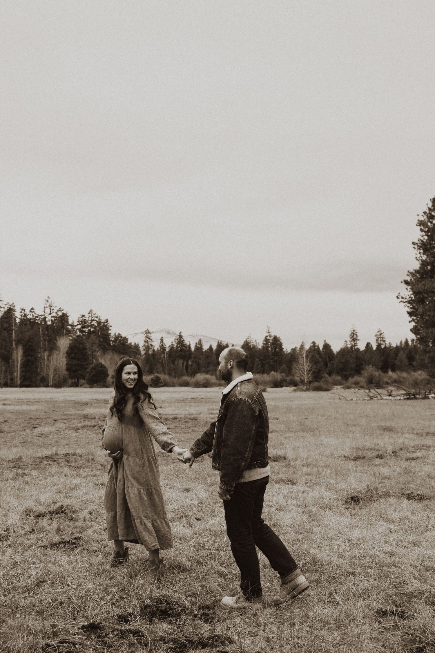 Pregnant woman in flowing dress leads partner by hand through an open field in a romantic maternity photo.